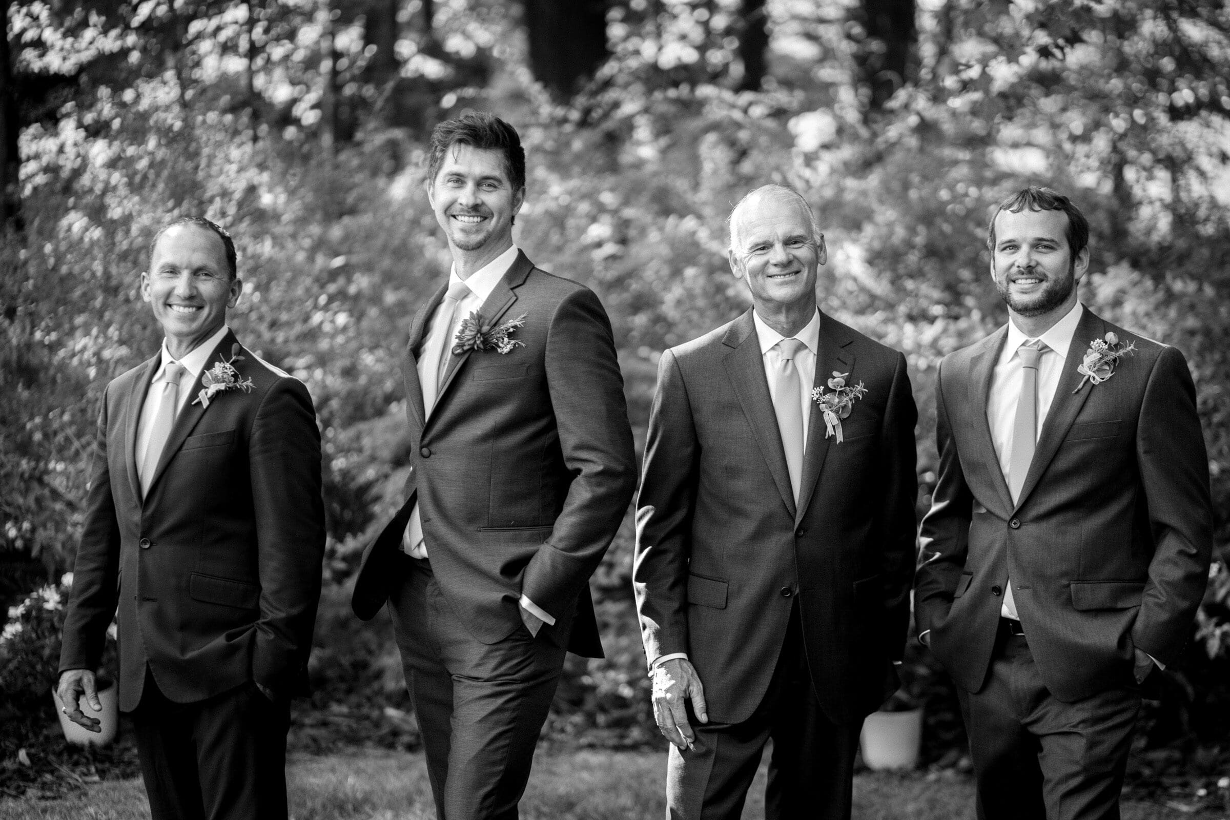 A black and white photo of four men standing outdoors in suits, smiling. The background is a lush garden with trees and shrubs. Each man has a boutonniere on their lapel.