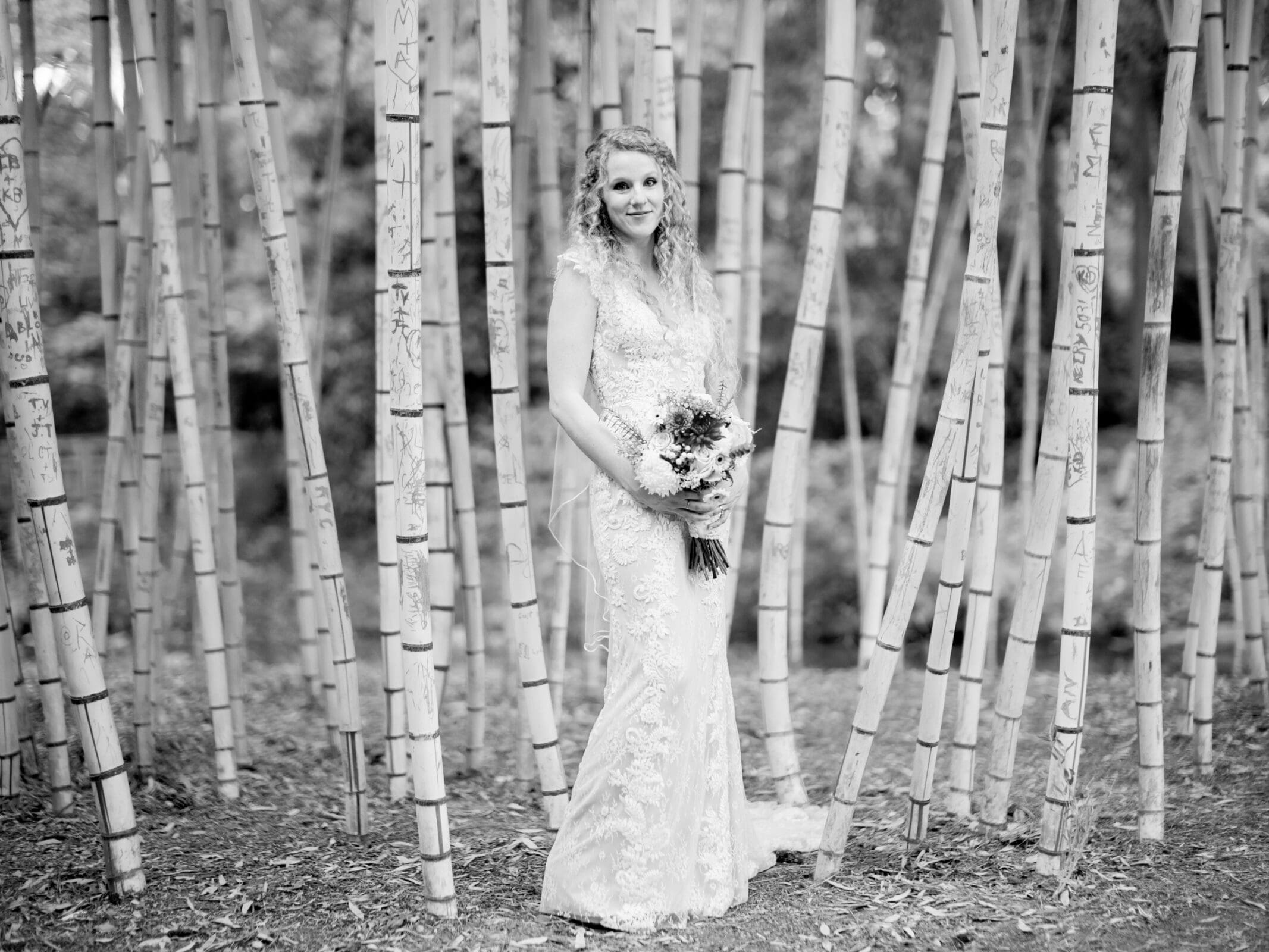 A bride in a lace wedding dress stands among tall bamboo stalks, holding a bouquet. The setting is serene and wooded, with carved initials visible on some bamboo stalks. The image is in black and white.