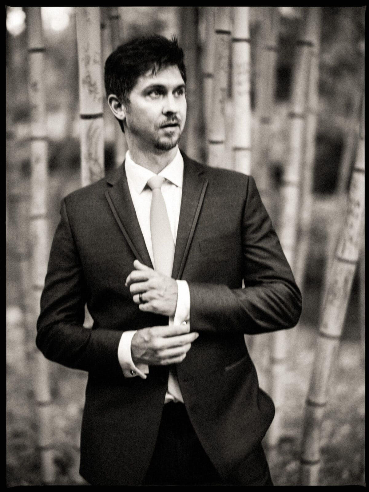 Black and white photo of a man in a suit standing in front of tall bamboo stalks. He is adjusting his jacket with a thoughtful expression, with a blurred outdoor background.
