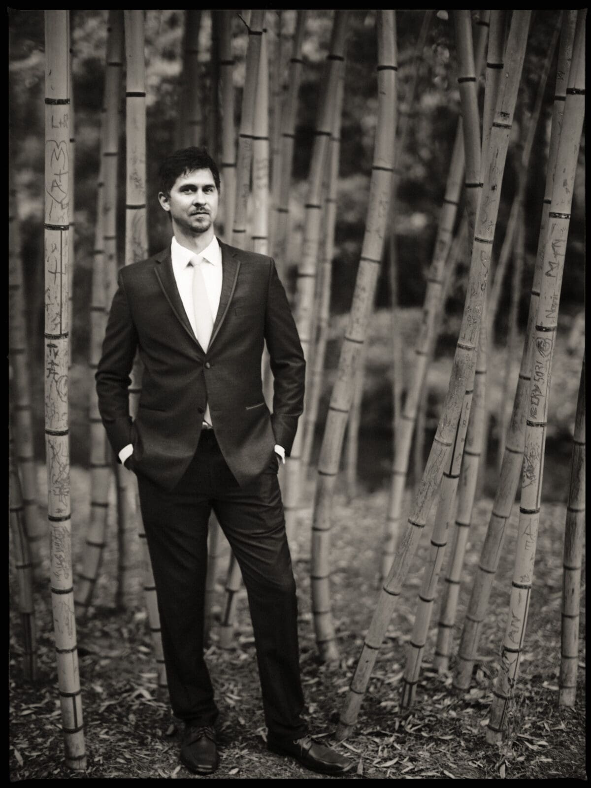 A man in a suit stands confidently among tall bamboo shoots. The black and white image creates a timeless, contemplative mood. He has his hands in his pockets and gazes thoughtfully into the distance.