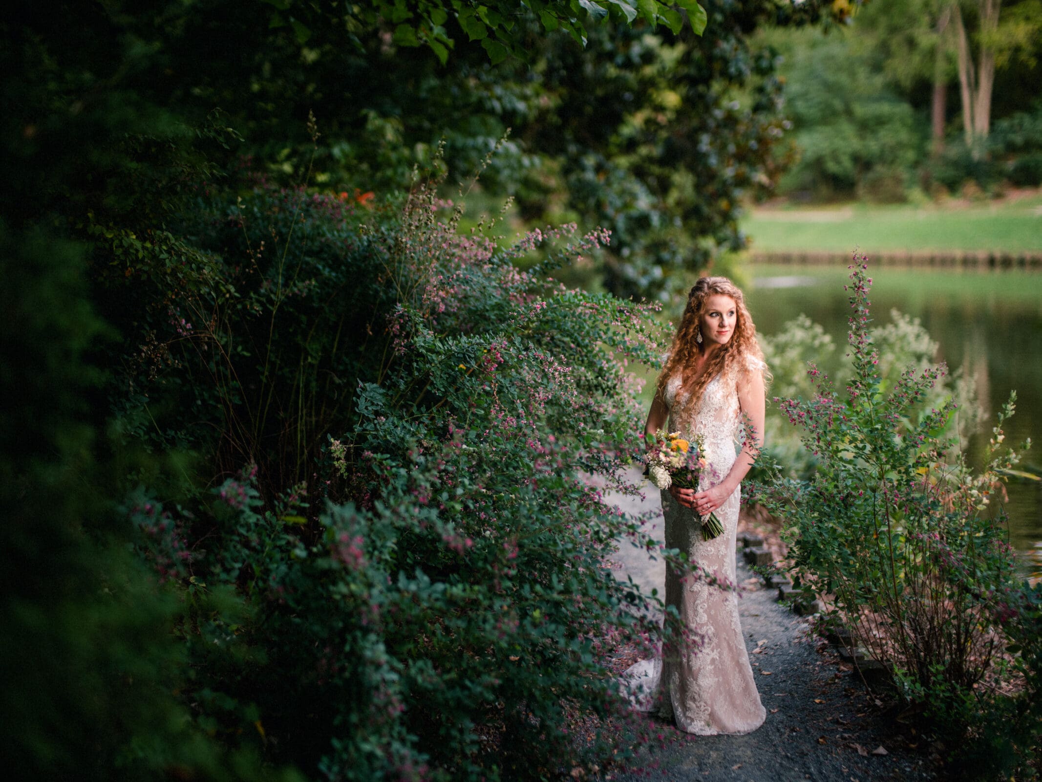 A woman in a floral gown stands on a garden path surrounded by lush greenery and small, flowering bushes. She holds a bouquet and looks thoughtfully to the side. A serene pond is visible in the background.