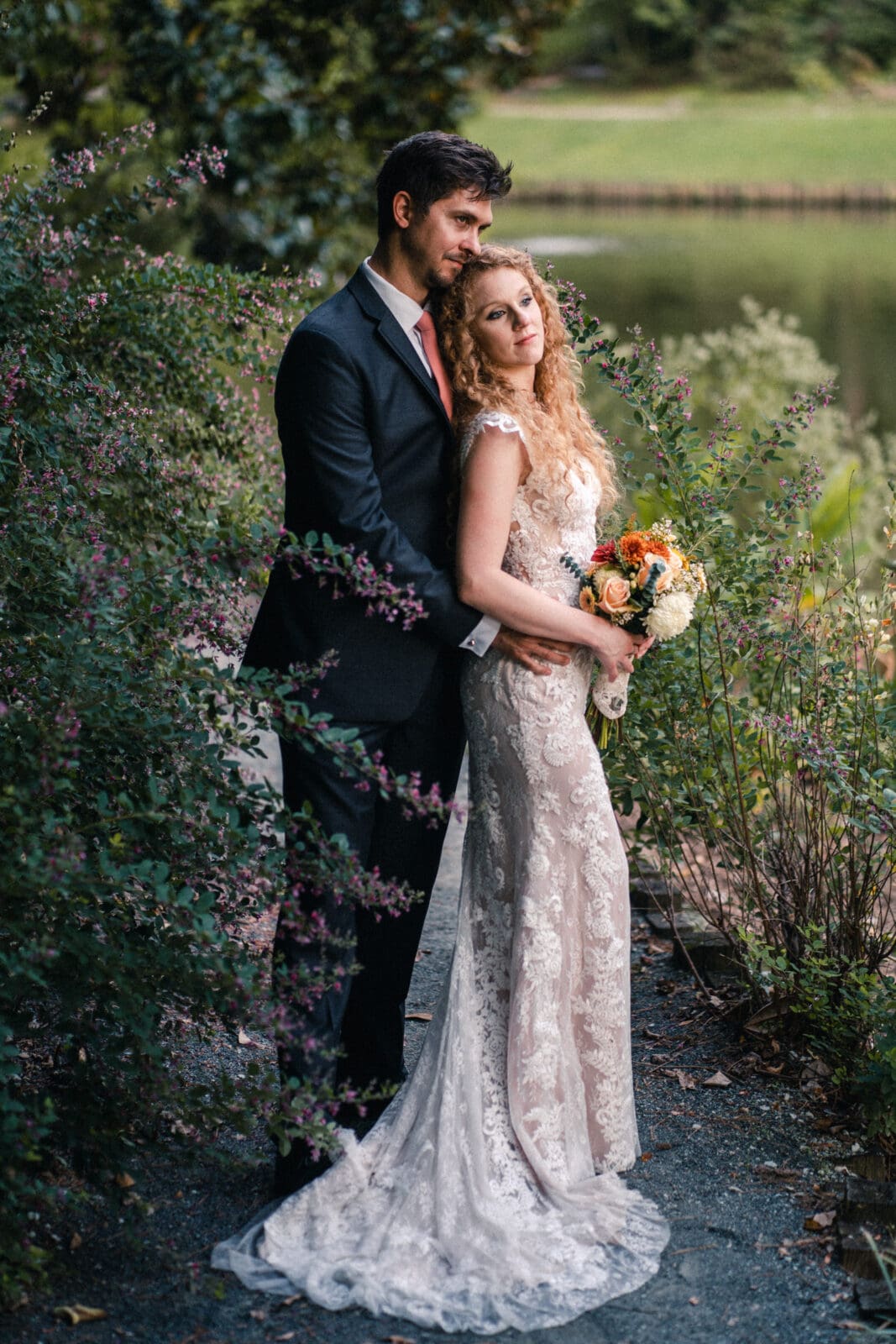 A couple stands in a lush garden by a pond. The woman, in a lace wedding dress, holds a bouquet of orange and white flowers. The man, dressed in a black suit with a red tie, lovingly embraces her from behind.