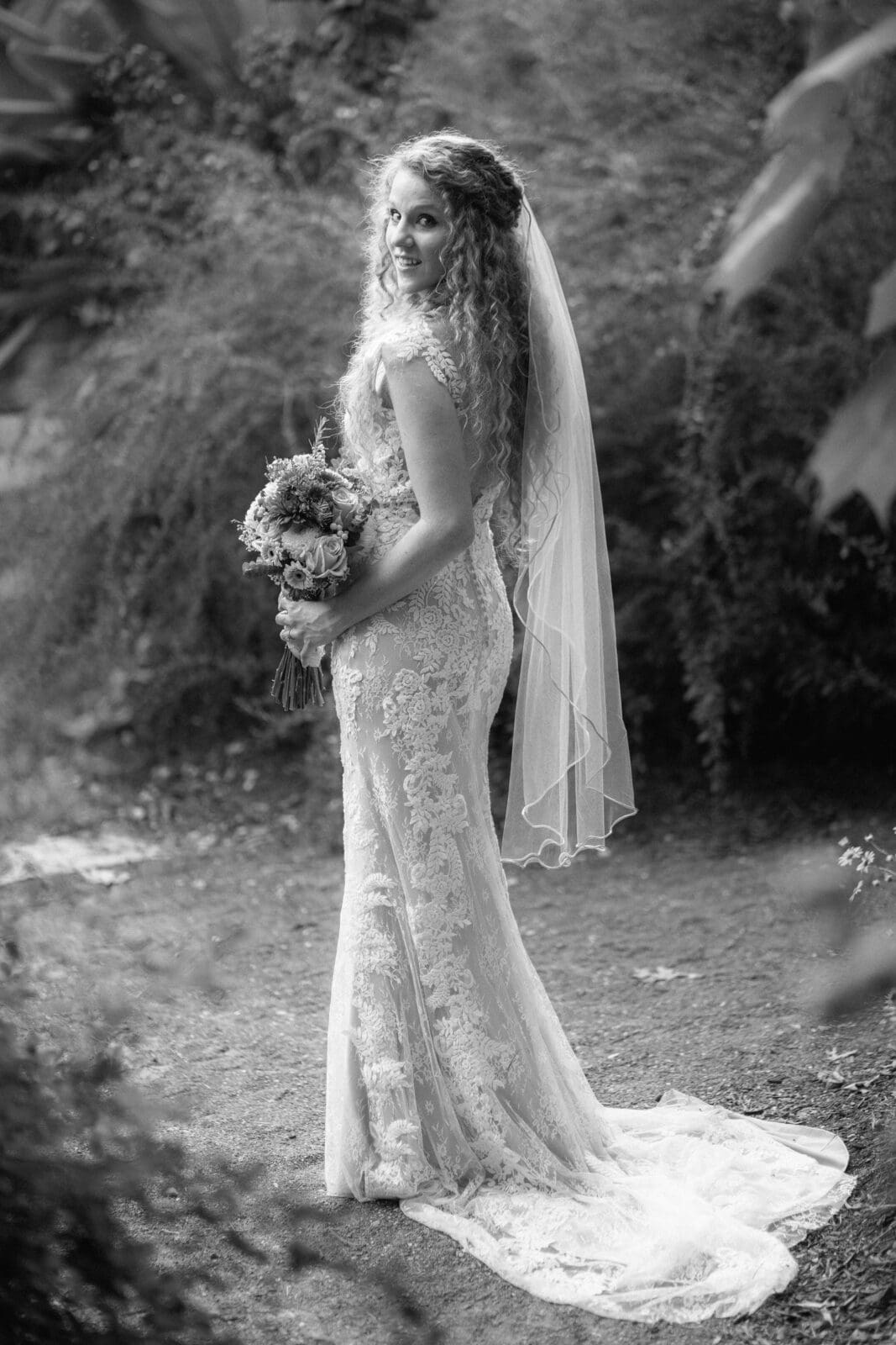 A bride with curly hair stands outdoors, wearing a lace wedding gown and veil, holding a bouquet of flowers. She smiles while looking over her shoulder amidst a lush, leafy backdrop. The image is in black and white.