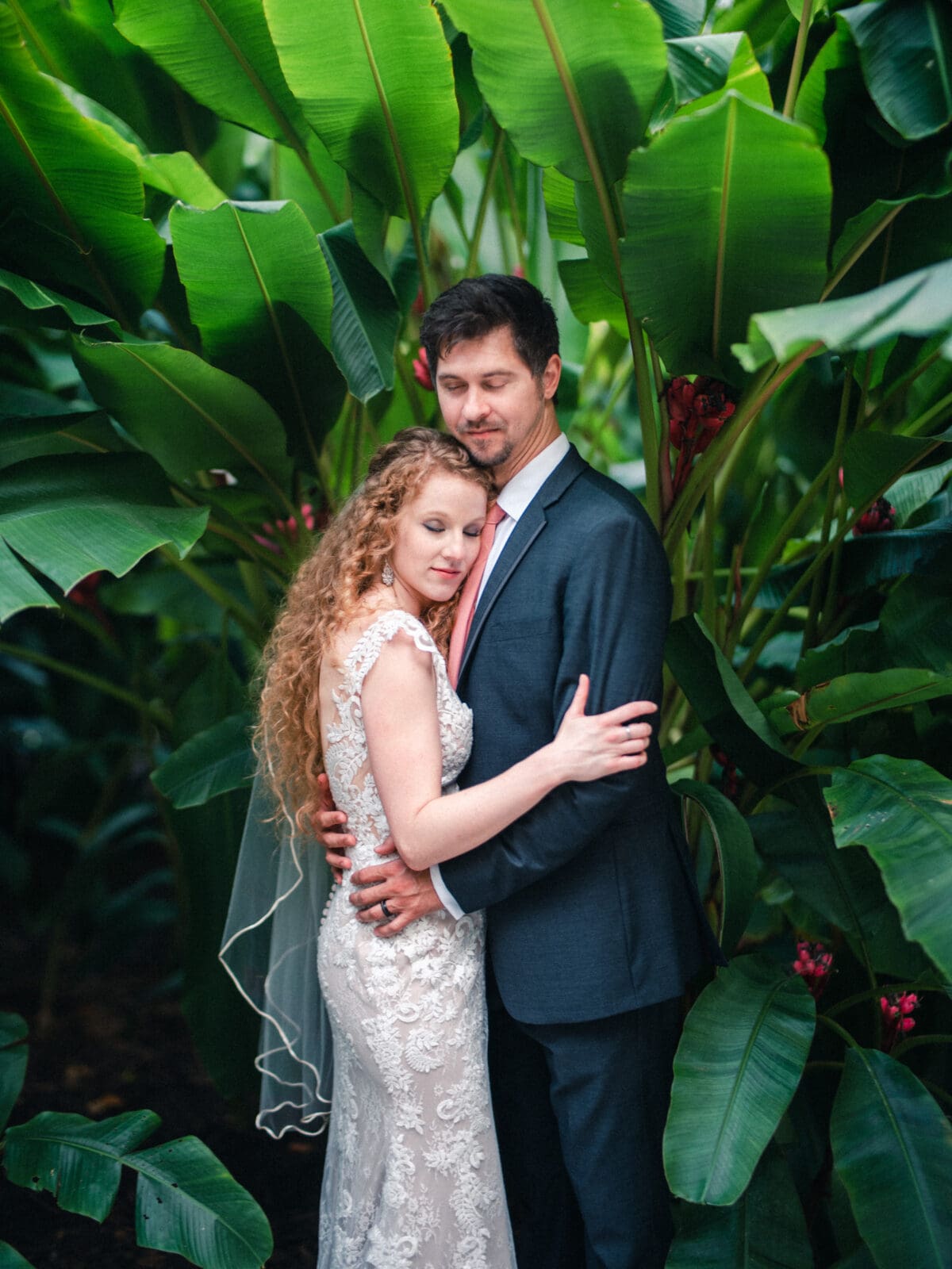 A couple embraces lovingly, surrounded by lush green tropical leaves. The woman is wearing a lace wedding dress, and the man is in a dark suit with a red tie. Both have their eyes closed, appearing peaceful and content.