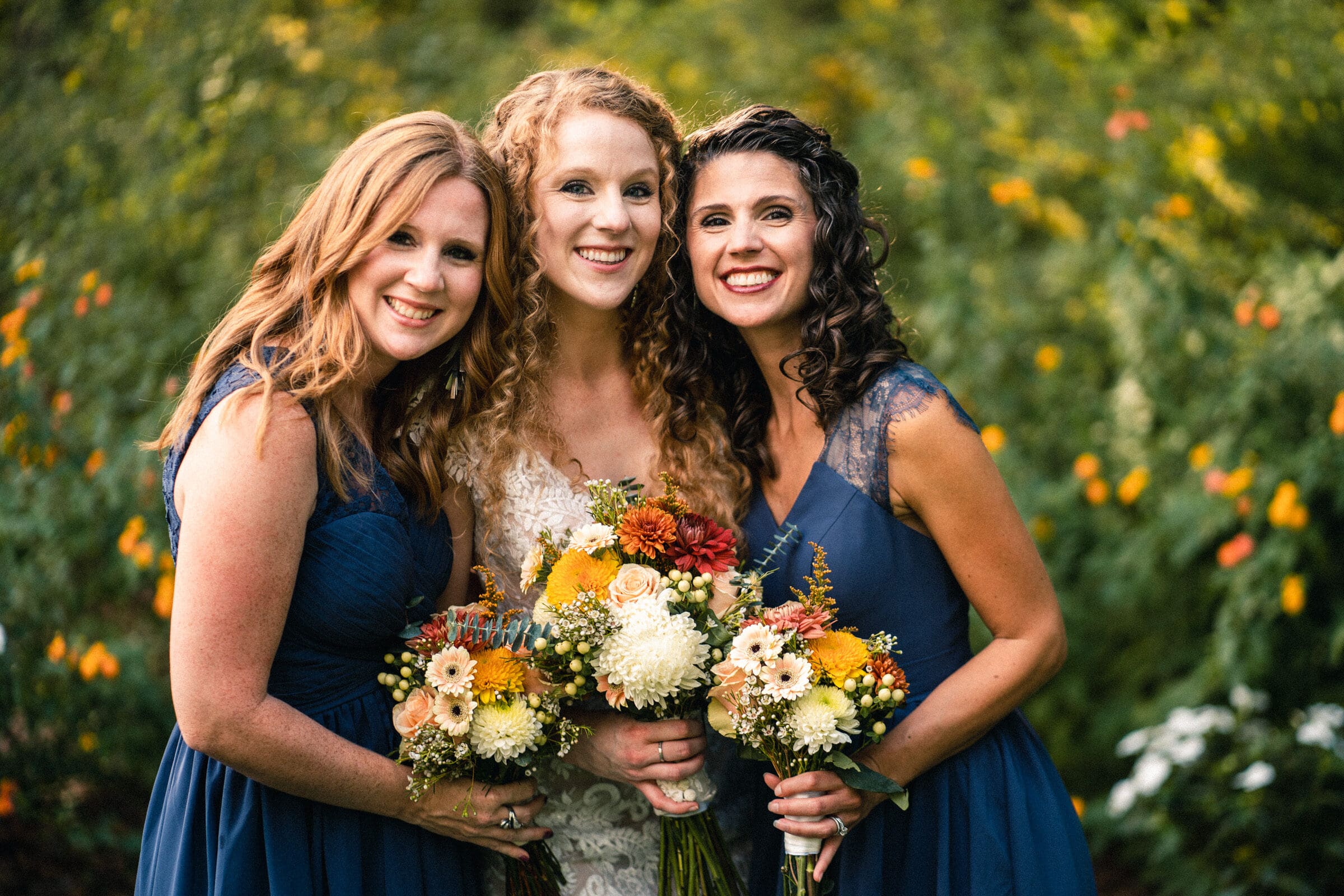 Three women in blue dresses are smiling together outdoors, holding bouquets of colorful flowers. The woman in the center is in a white gown. The background is lush and green, suggesting a garden setting.