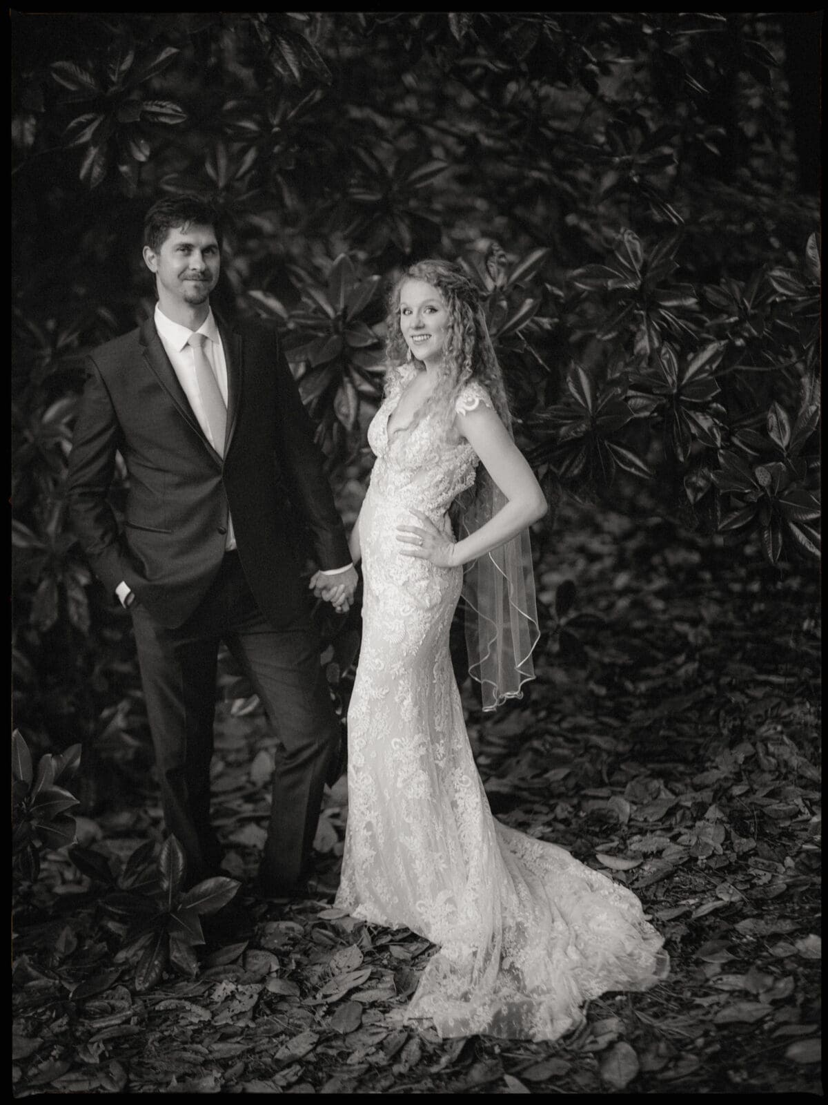 A black and white photo of a bride and groom standing outdoors. The bride wears a lace gown with a veil, and the groom is in a dark suit with a tie. They are holding hands against a backdrop of large leafy plants.