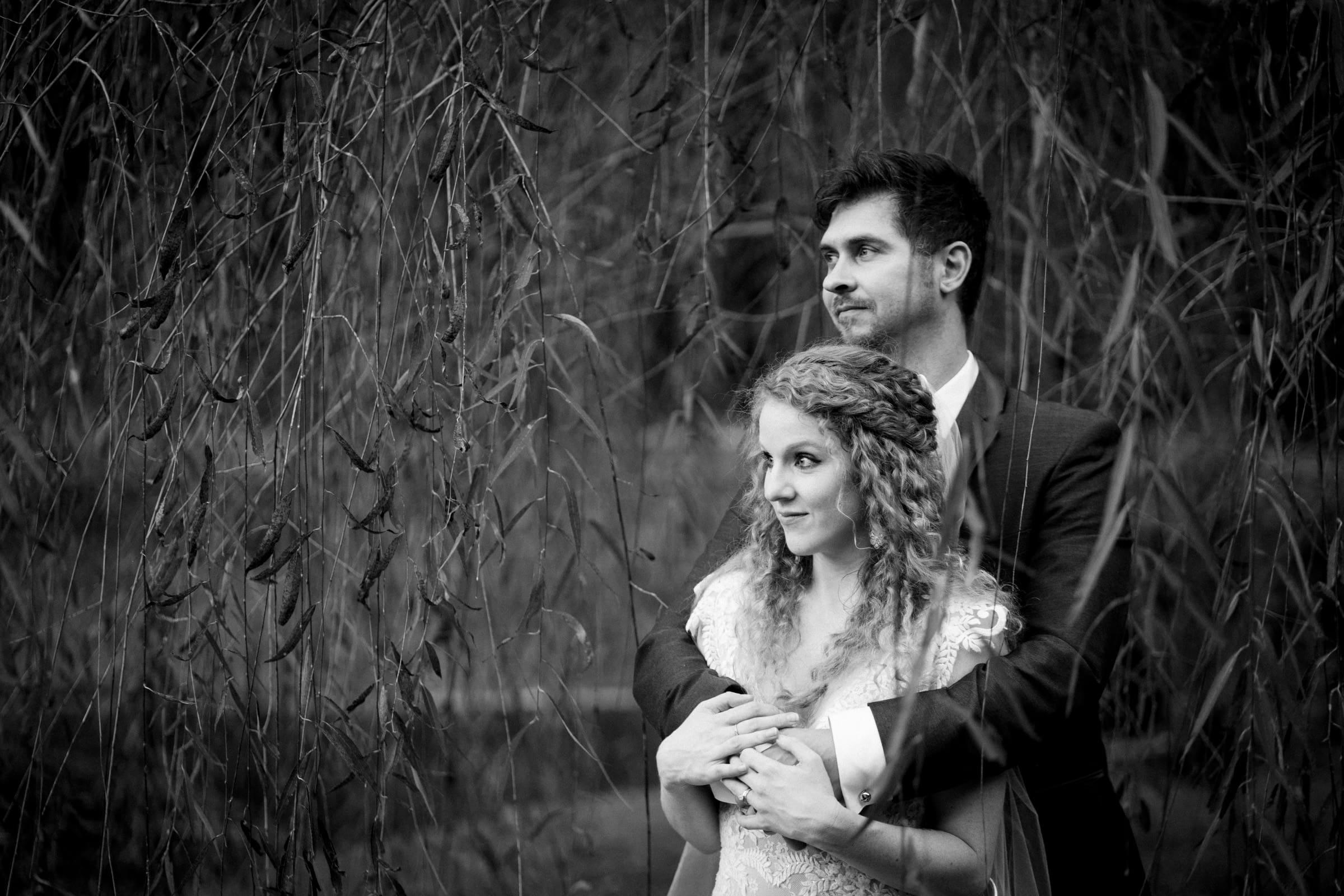 A black and white photo of a couple in formal attire. The man stands behind the woman, arms around her waist. They are surrounded by hanging branches, creating a romantic and serene atmosphere.