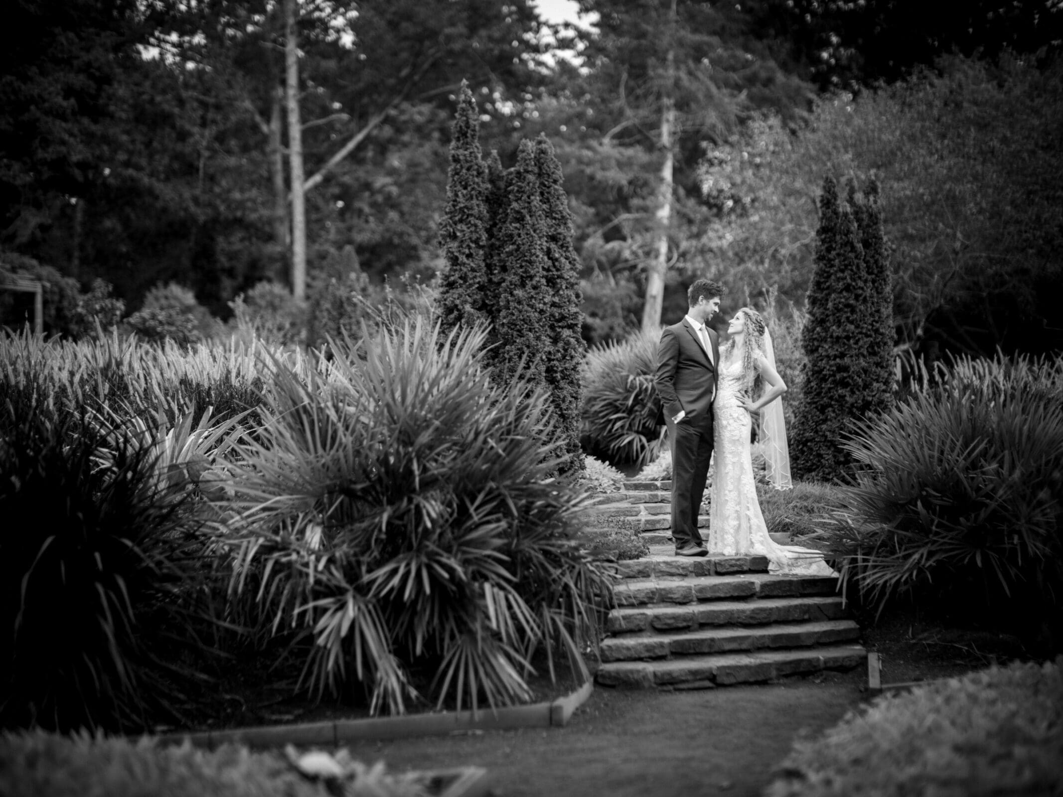 A bride and groom share a moment on a stone pathway surrounded by lush greenery and tall trees in a garden setting. Black and white photo captures the couple in a tender embrace.