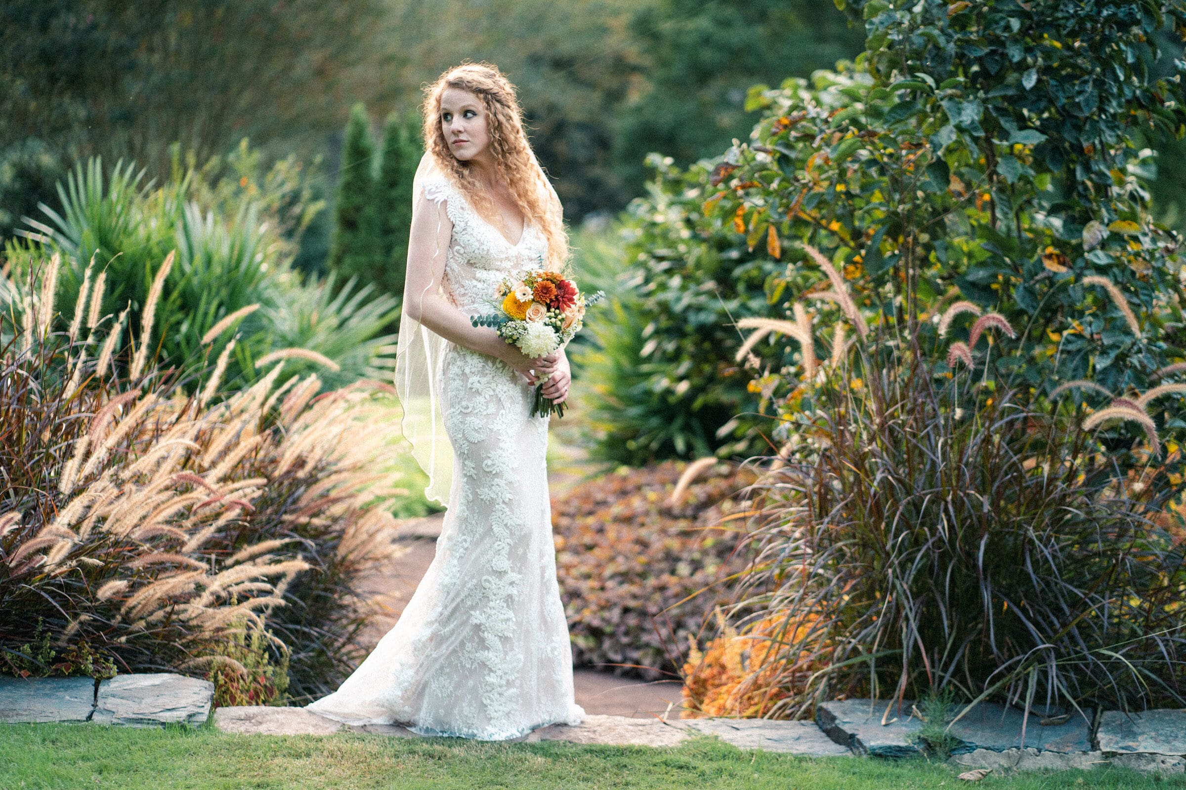 A bride in a lace wedding dress holds a bouquet while standing in a lush garden. She gazes to the side, surrounded by greenery and ornamental grasses under soft, natural light.