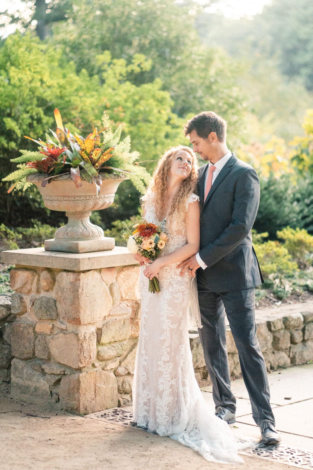 A bride and groom stand together in a garden setting. The bride is in a lace dress holding a bouquet, while the groom, in a suit, stands beside her with an arm around her waist. They are near a stone planter with colorful flowers and greenery.