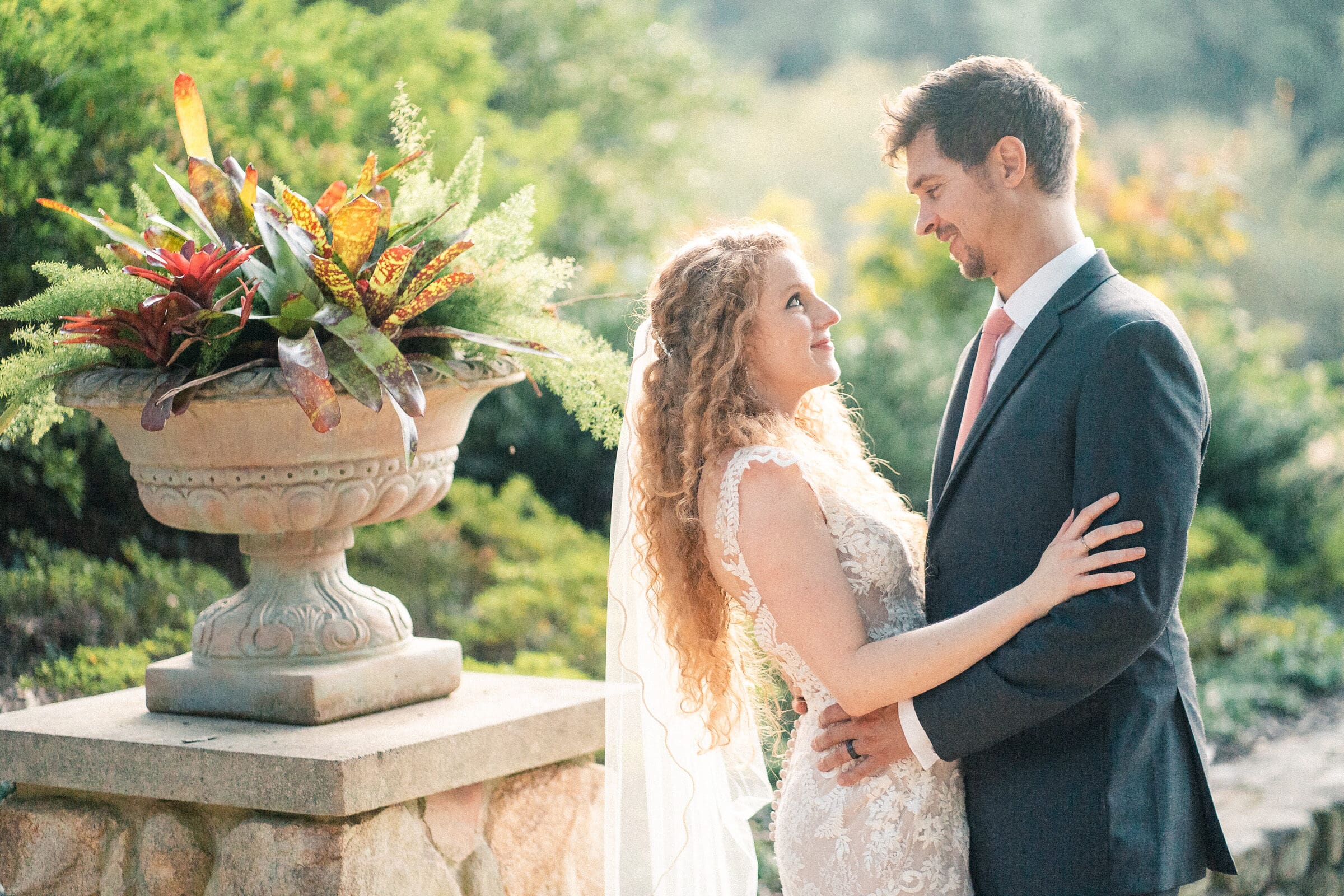 A bride and groom gaze at each other lovingly. The bride, in a lace gown and veil, and the groom, in a dark suit, stand next to a stone planter with vibrant foliage. The background is a lush, sunlit garden.
