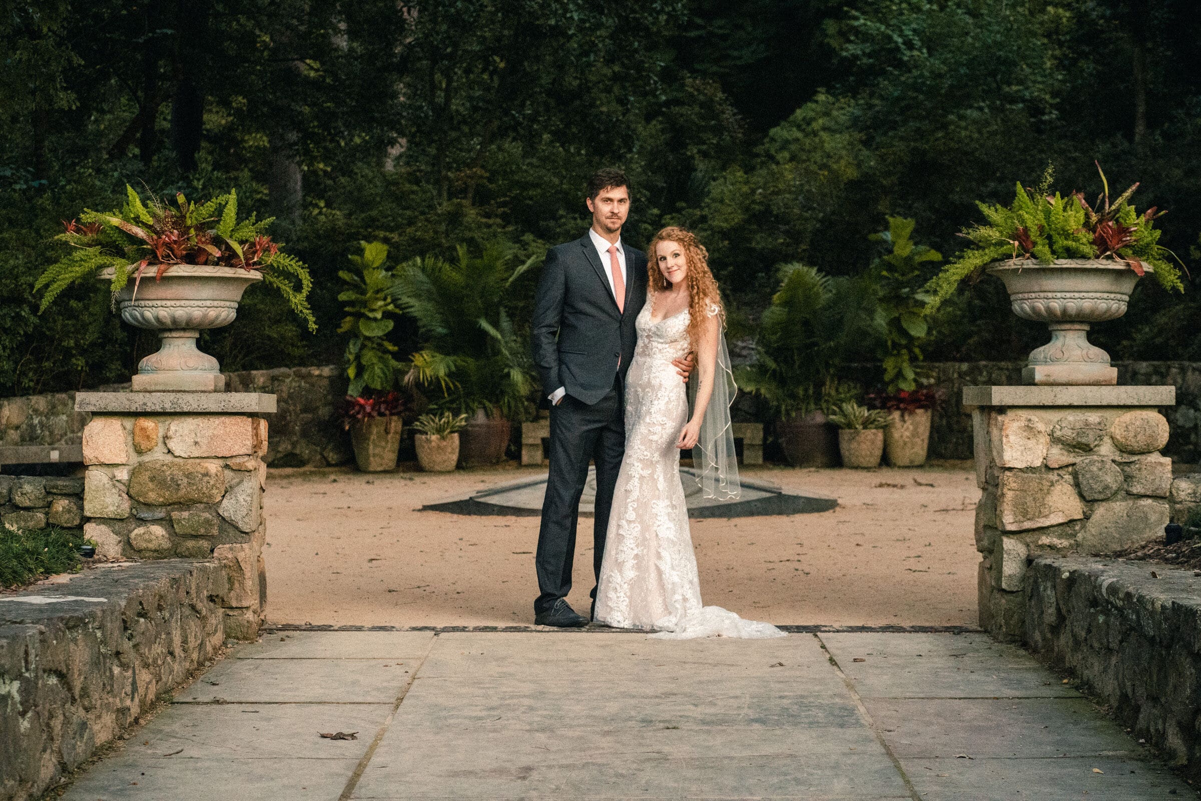 A bride and groom stand in a garden setting with potted plants and stone planters. The groom wears a dark suit, while the bride is in a white lace gown with a veil. They pose on a stone pathway surrounded by lush greenery.