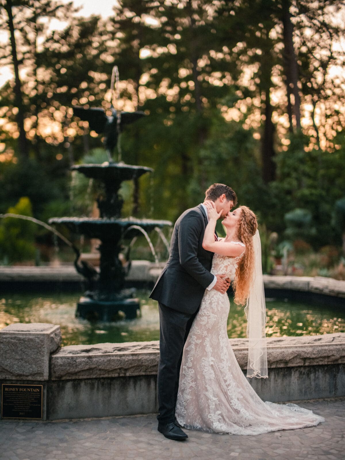 A bride and groom share a romantic kiss in front of a fountain at sunset. The bride wears a lace gown with a veil, and the groom is dressed in a dark suit. The setting includes lush greenery and a serene atmosphere.