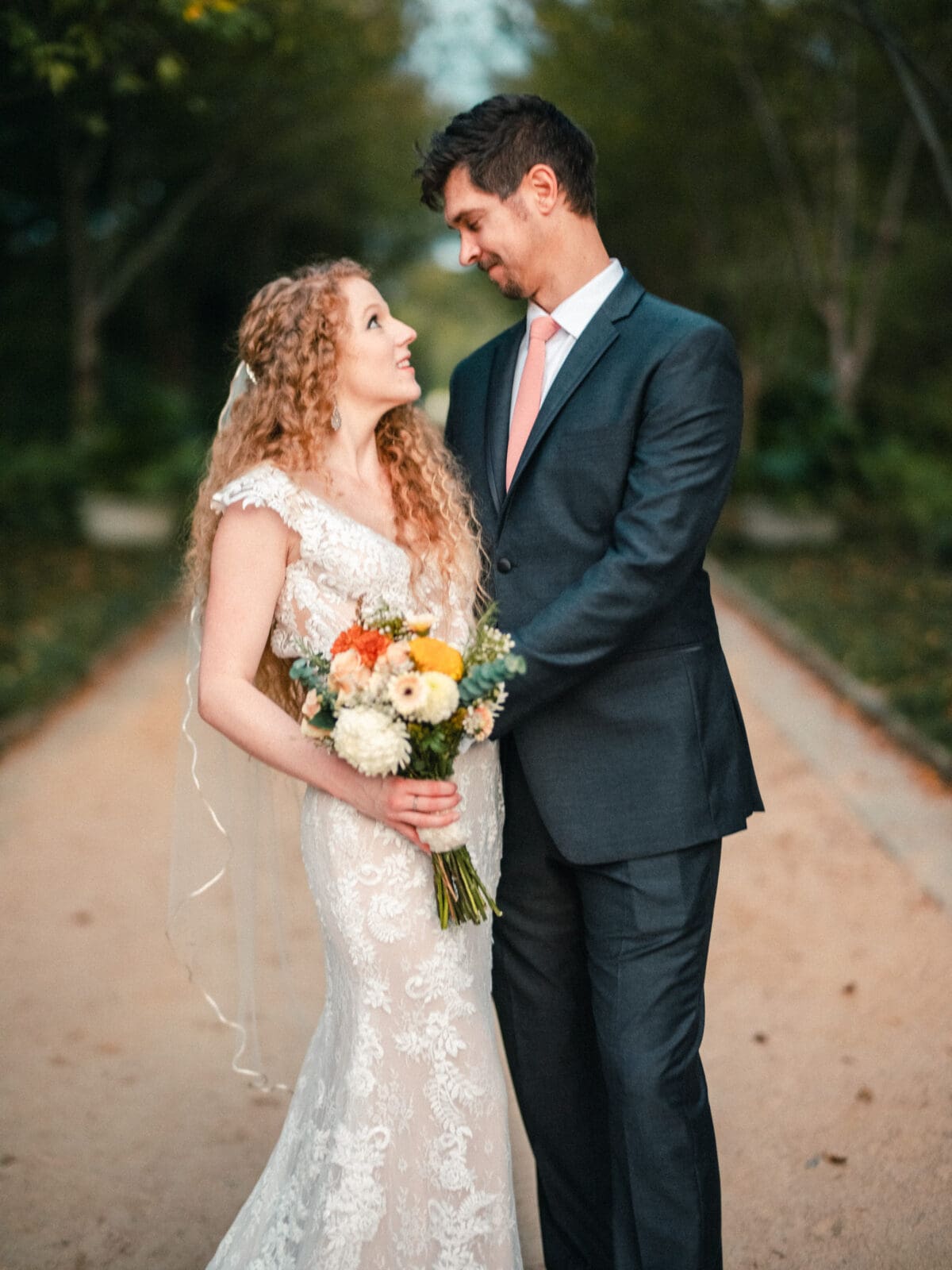 A bride and groom stand on a path surrounded by greenery. The bride, in a lace wedding dress, holds a bouquet of flowers and gazes up at the groom, who is wearing a suit and pink tie. Both are smiling at each other.