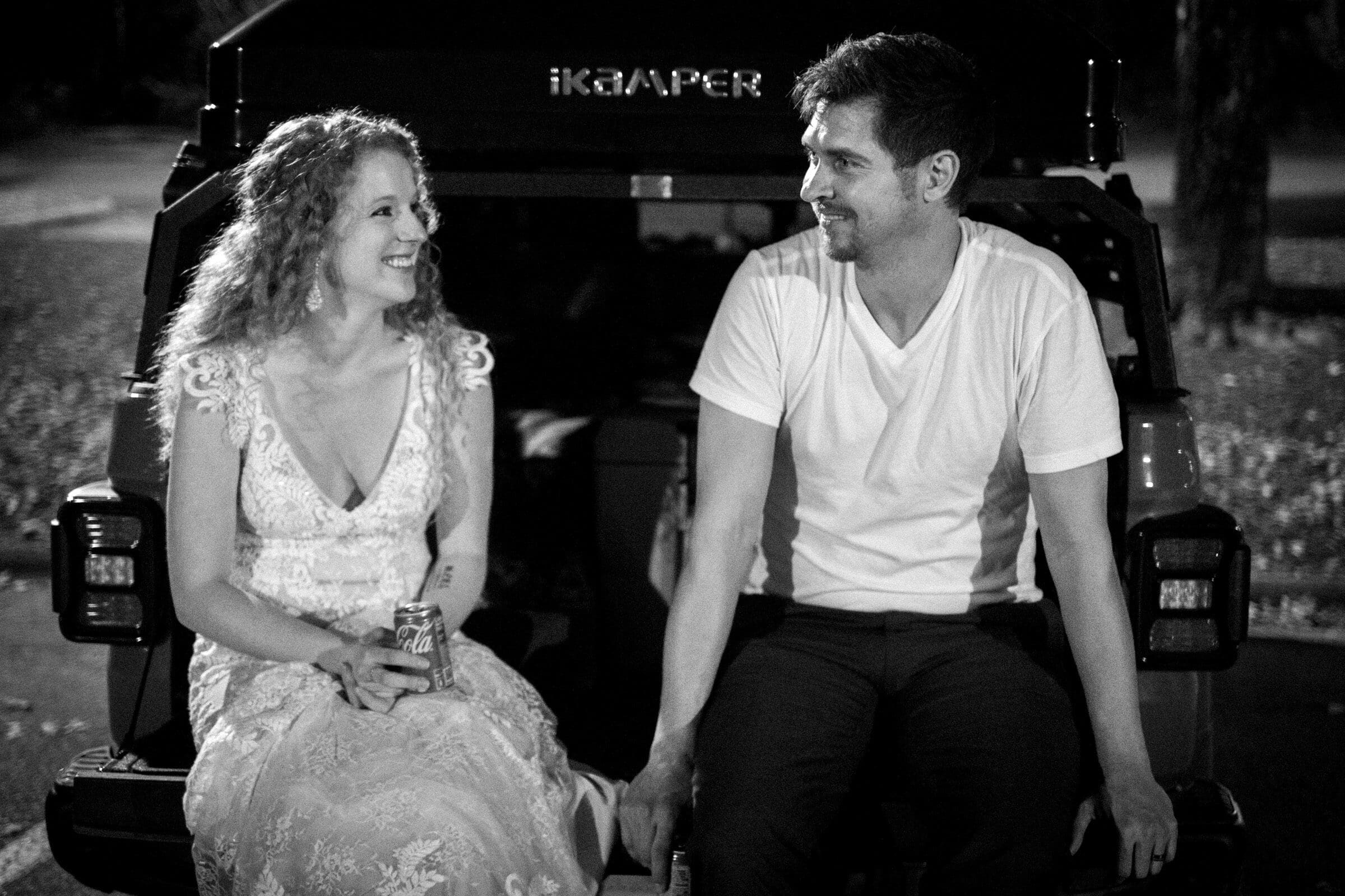 A woman in a lace dress and a man in a white t-shirt sit on the open tailgate of a vehicle at night. Both are smiling and holding drinks, with soft lighting creating a relaxed atmosphere.