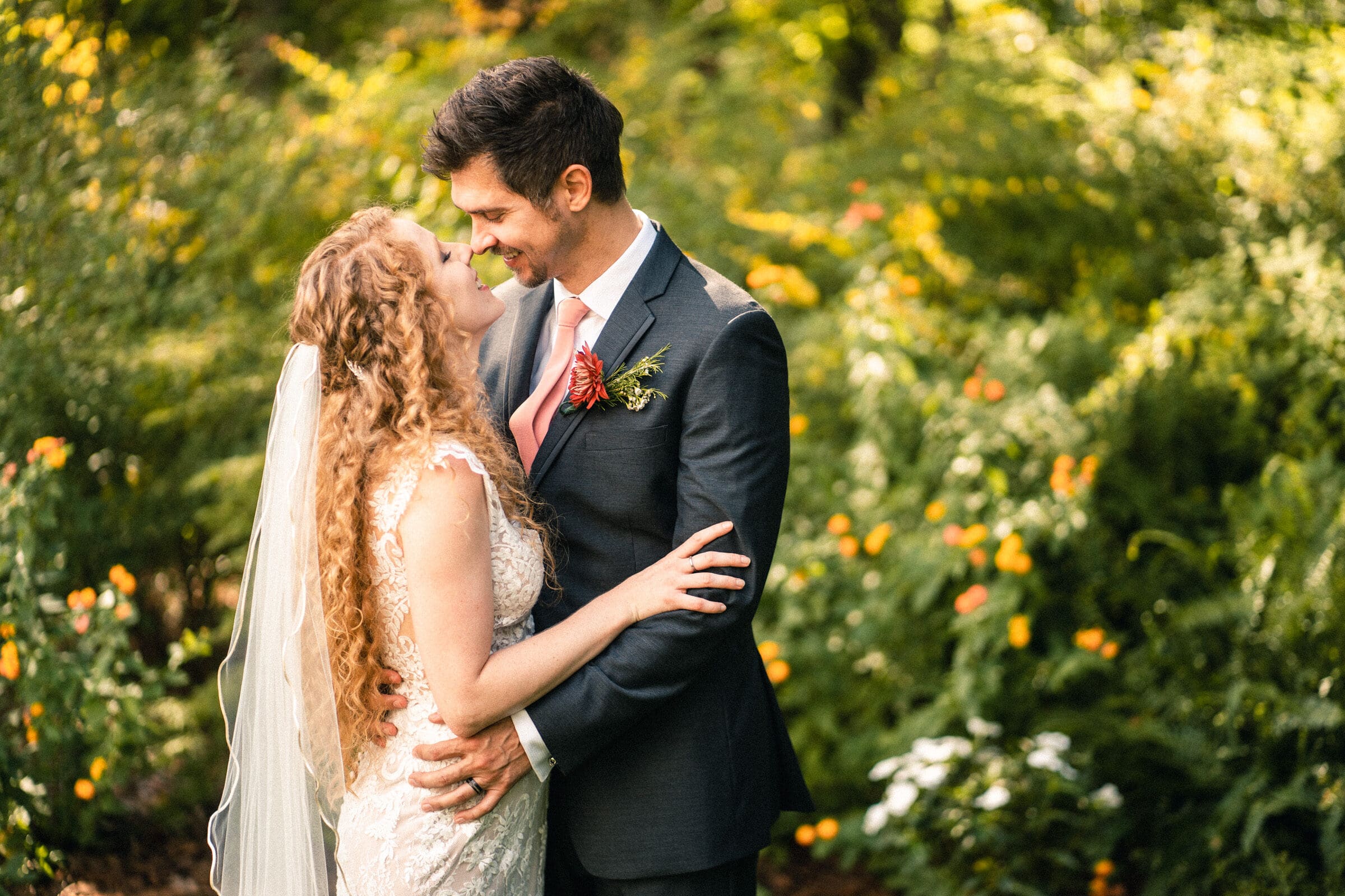 A bride in a lace gown and veil embraces a groom in a dark suit with a pink tie and boutonniere. They stand in a lush garden with vibrant flowers, smiling at each other affectionately.
