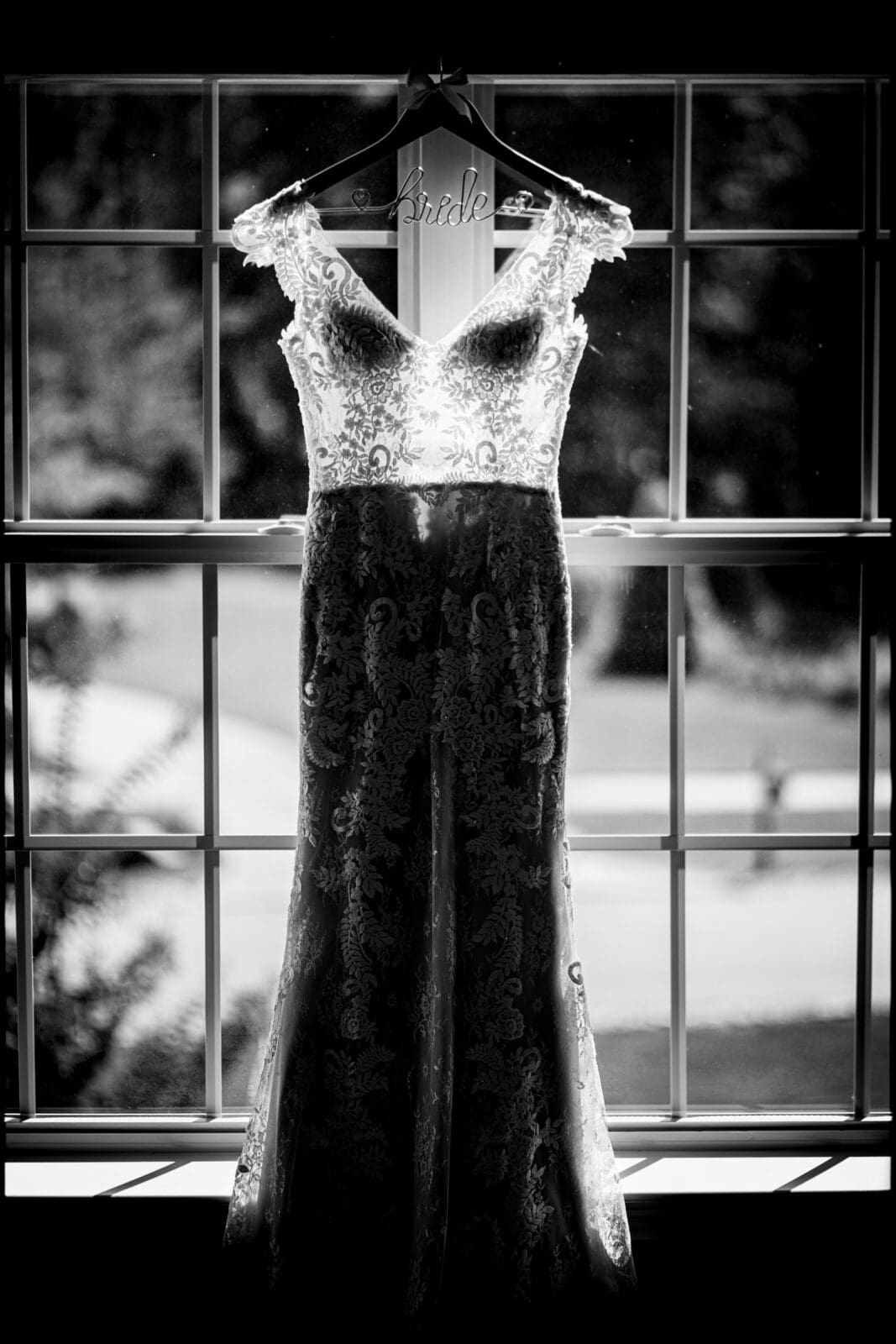 A black and white photo of an elegant lace wedding dress hanging on a wooden hanger in front of a window. Sunlight filters through the window, highlighting the intricate patterns of the dress. The background shows a blurred outdoor scene.