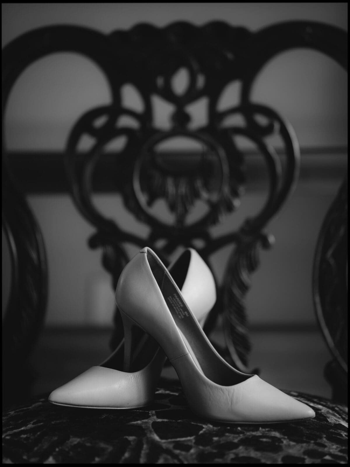 A pair of elegant high-heeled shoes is artistically arranged and placed on a luxurious, ornately carved chair. The black and white image highlights the intricate patterns and textures, creating a vintage and sophisticated ambiance.