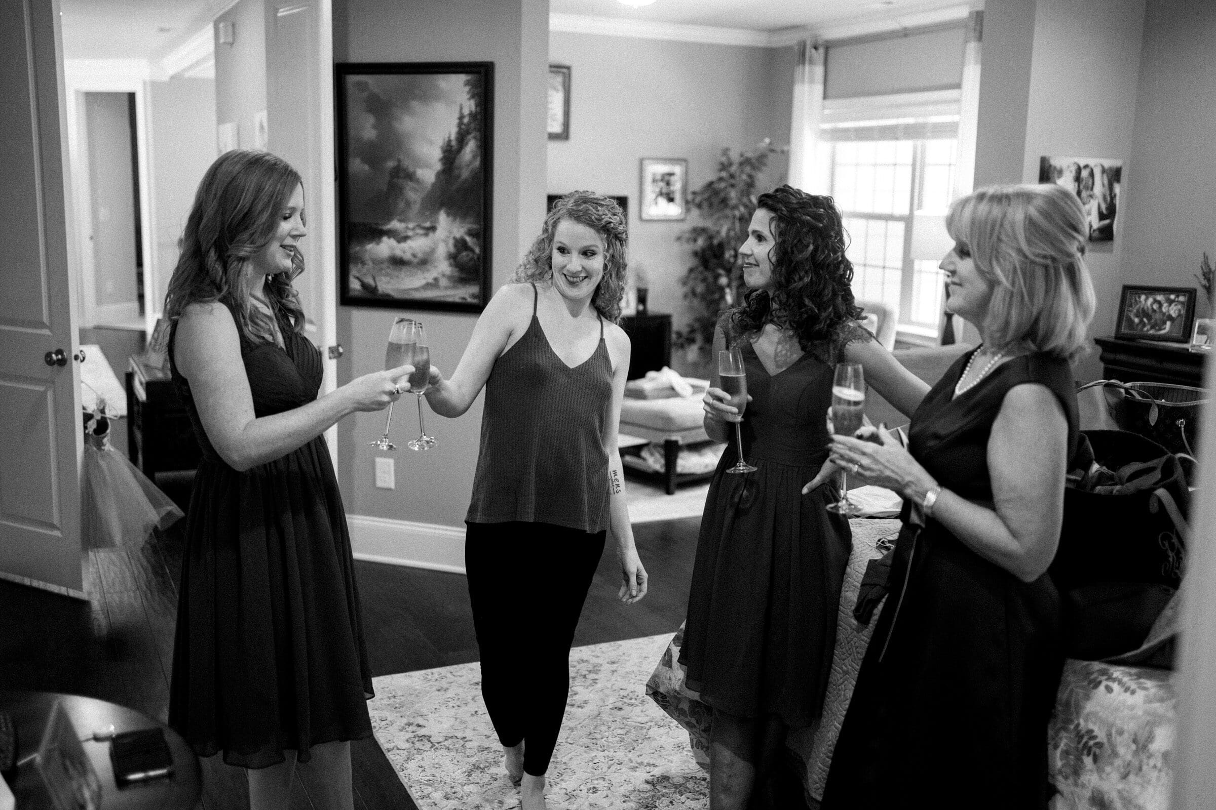 Four women in a living room, dressed in formal and casual attire, smile and hold champagne glasses. They appear to be celebrating or enjoying a special occasion together. The room is elegantly decorated with framed art and large windows.