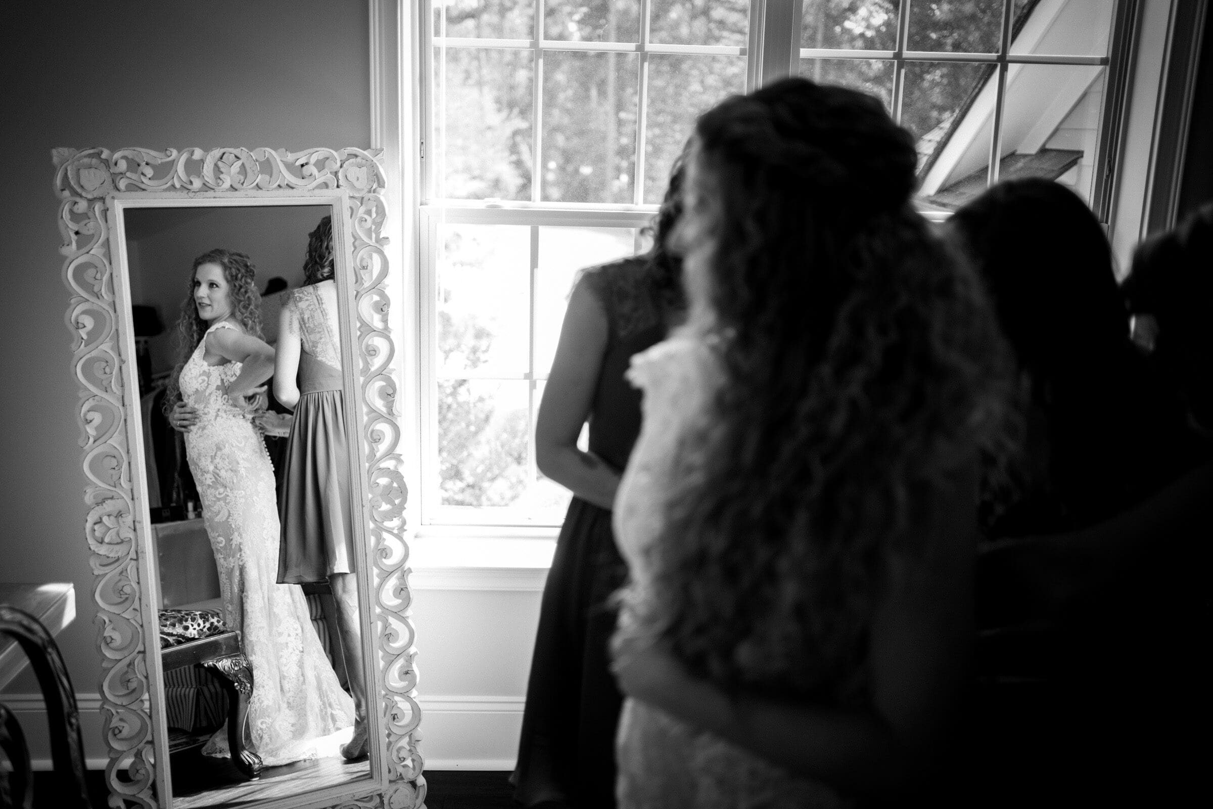 A bride in a lace wedding dress looks at her reflection in an ornate mirror. She stands in a room with large windows, surrounded by blurred figures, who appear to be assisting her. The scene captures a moment of preparation. Black and white image.
