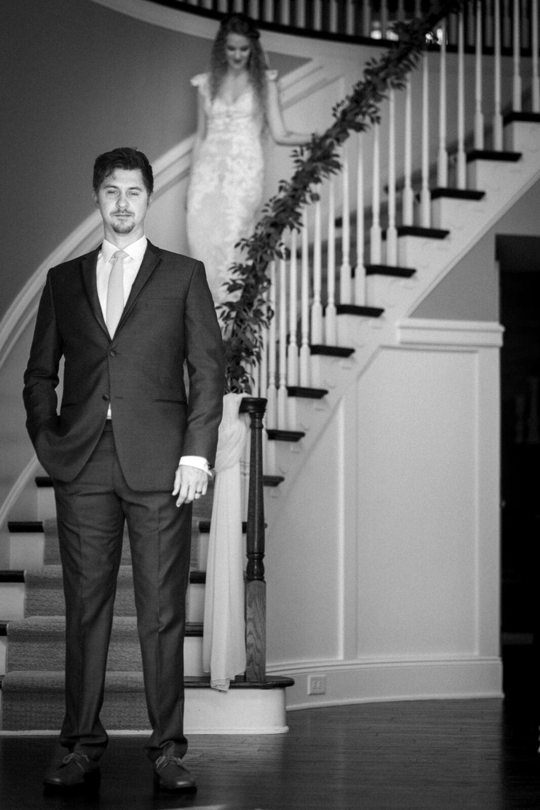 A black and white image of a man in a suit standing at the bottom of a staircase, with a woman in a wedding dress descending the stairs behind him. The staircase is decorated with greenery.