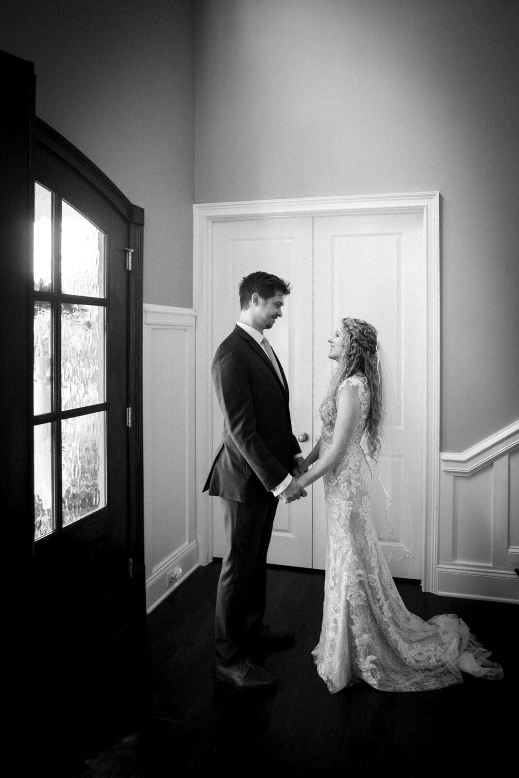 A black and white photo of a bride and groom standing indoors, holding hands and facing each other. The bride wears a long, lace wedding dress, and the groom is in a suit. They are by a door with glass panels, near a stairway.