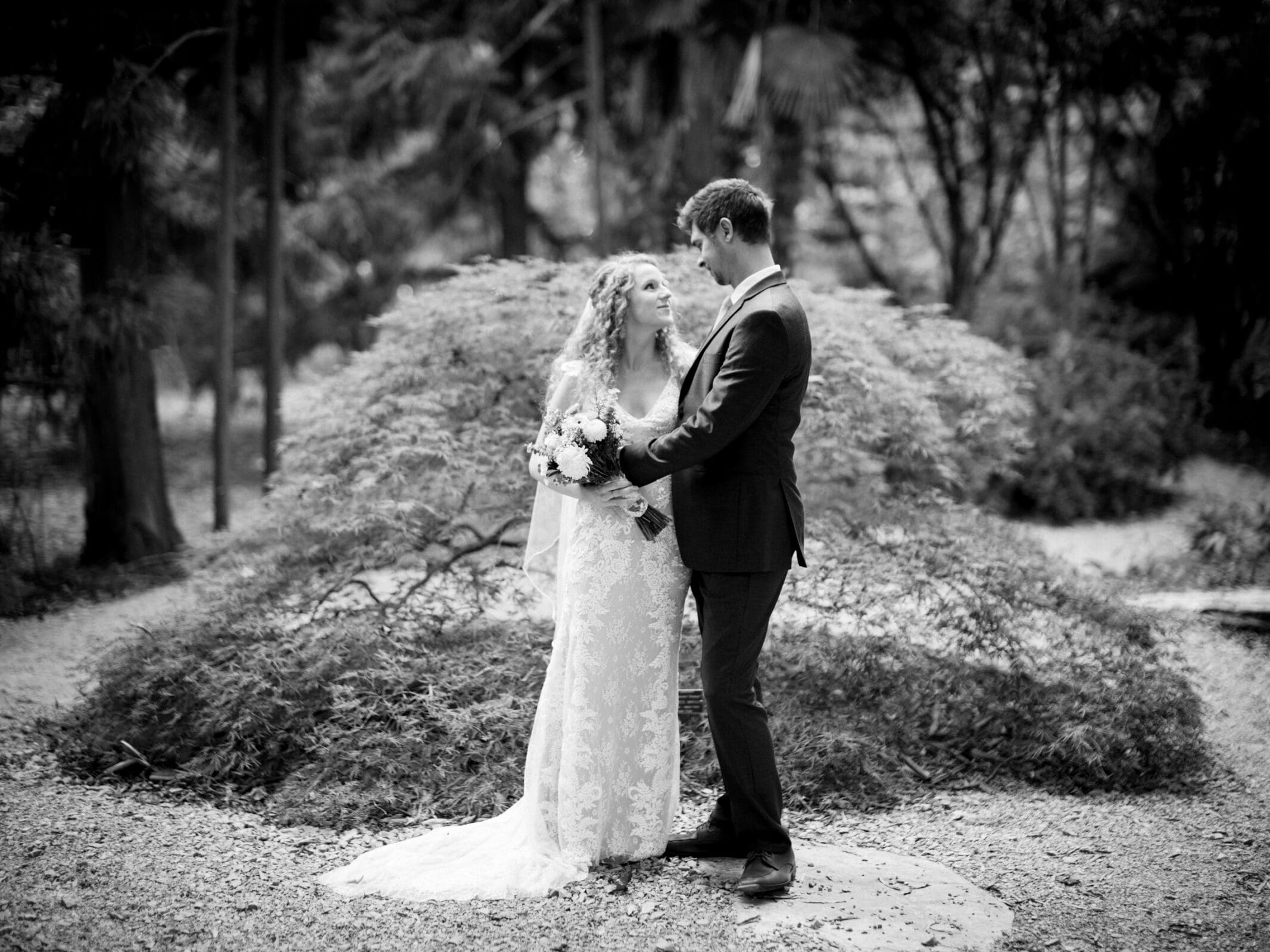 A bride and groom stand facing each other in a garden setting. The bride is in a lace dress, holding a bouquet, while the groom wears a dark suit. They are surrounded by lush greenery, creating a serene and romantic atmosphere.