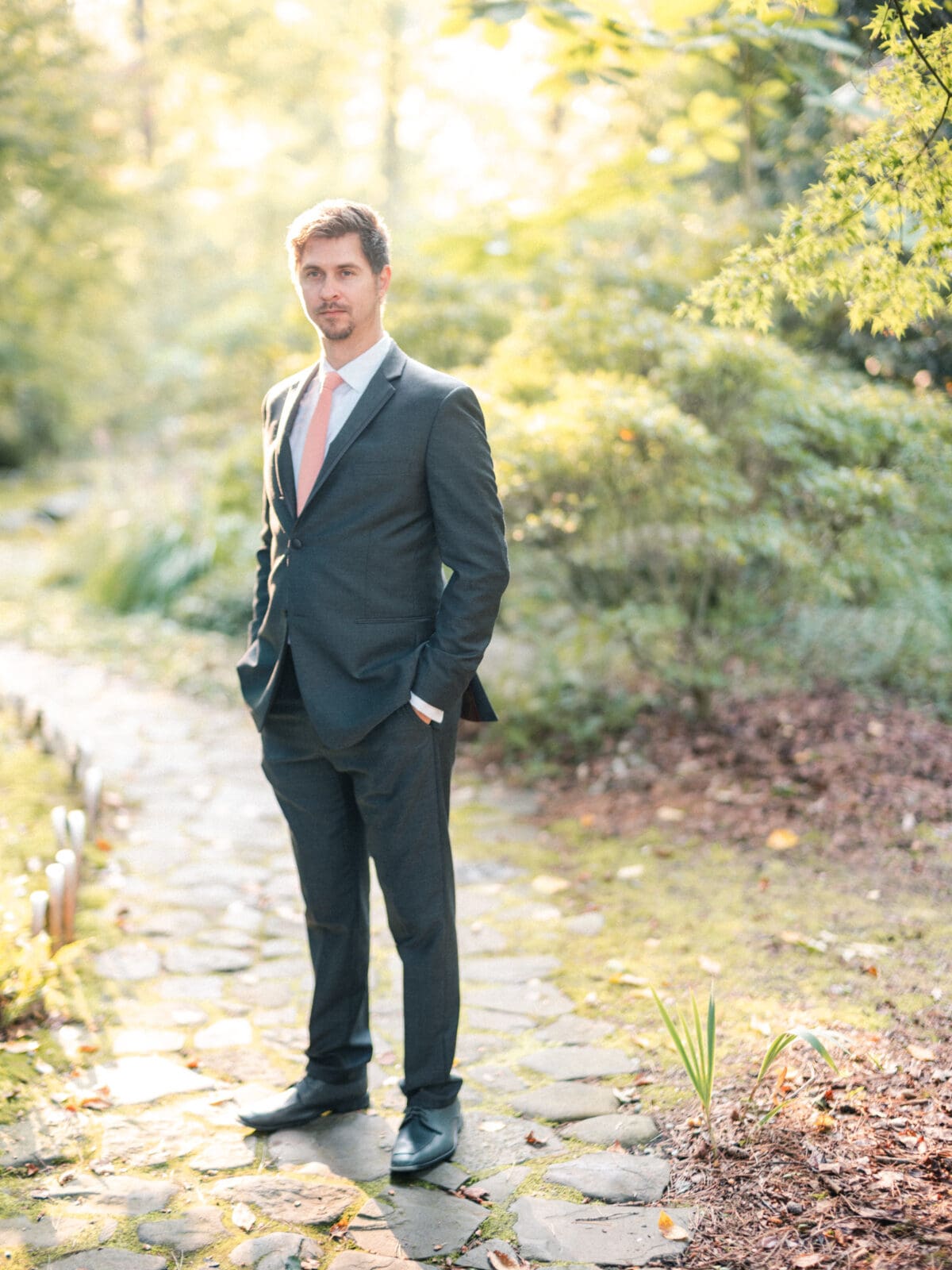 A man in a suit with a light pink tie stands on a stone path surrounded by lush greenery and sunlight filtering through the trees.