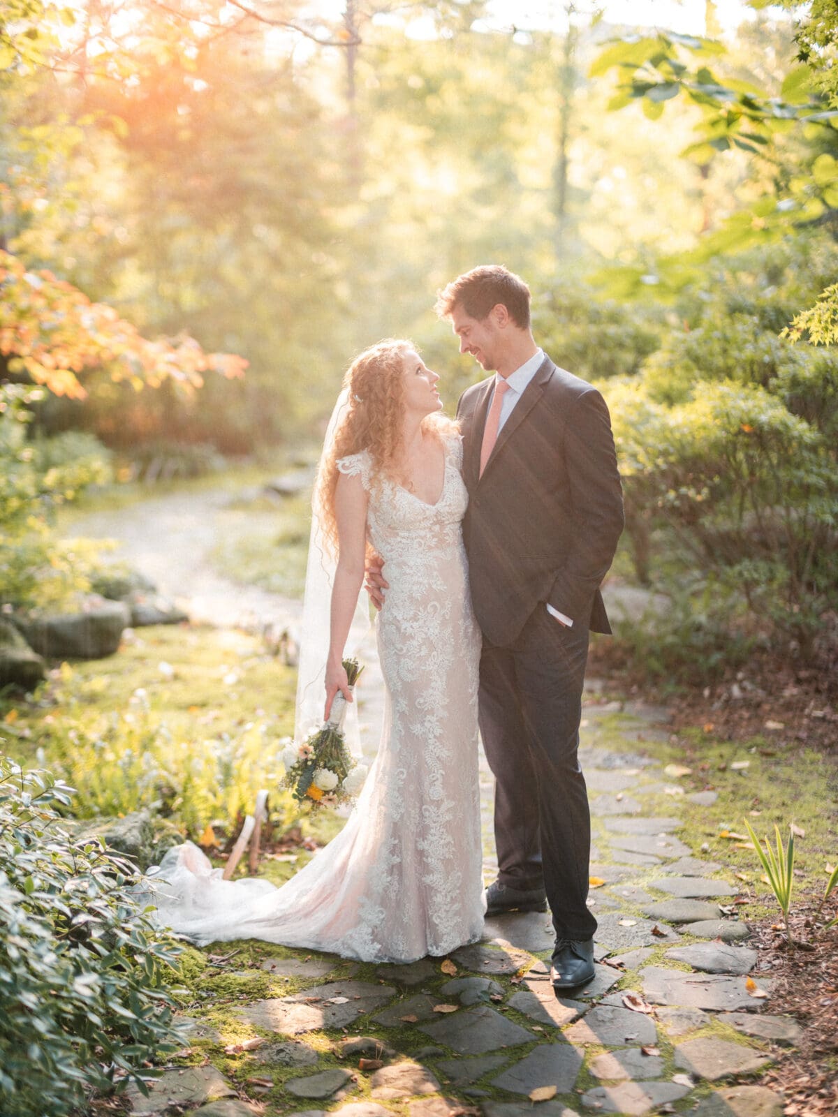 A bride and groom stand on a stone path in a sunlit garden. The bride wears a lace gown and veil, and holds a bouquet. The groom is in a suit with a pink tie. They smile at each other, surrounded by lush greenery and warm sunlight.