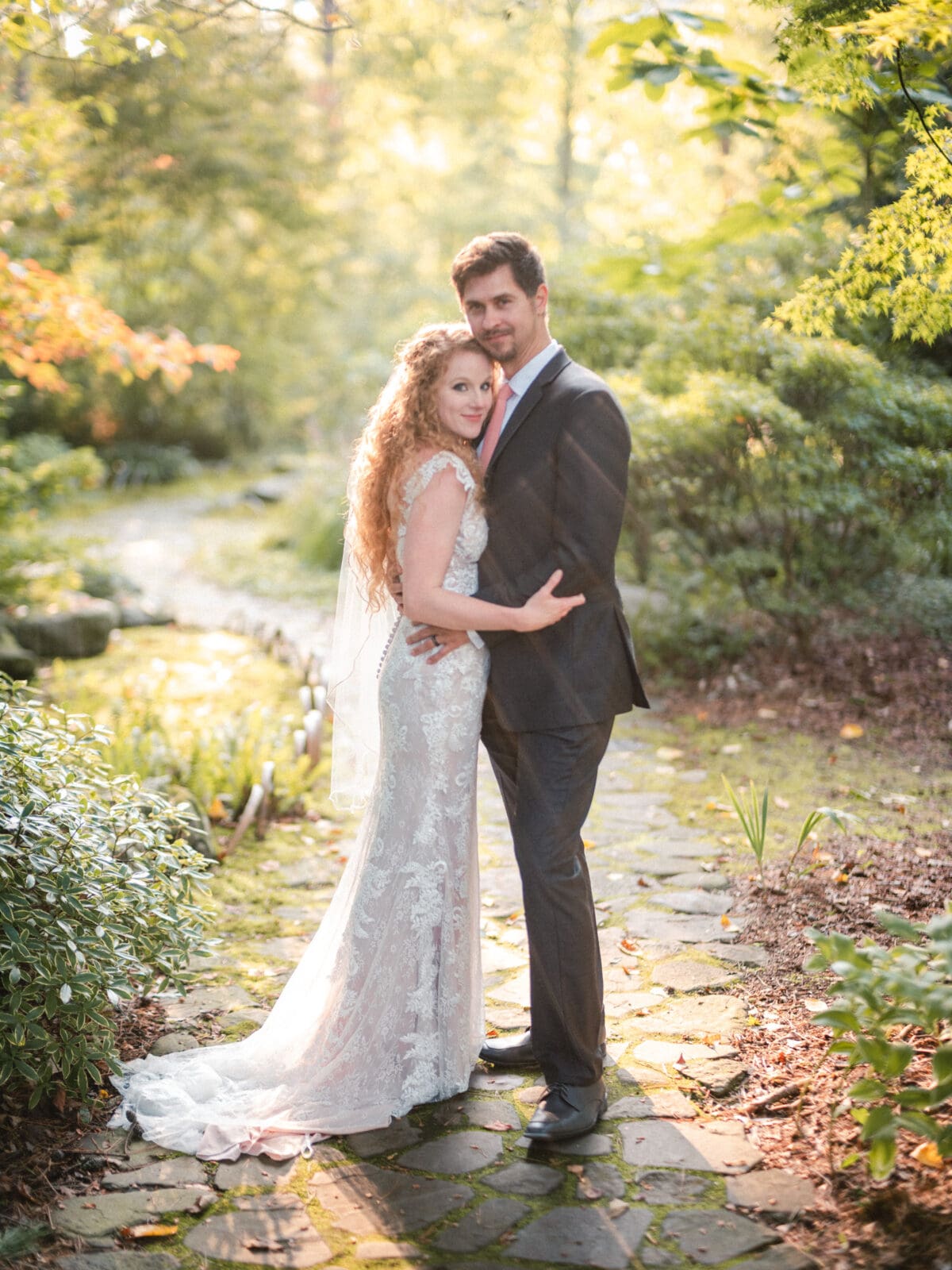 A couple stands on a stone path in a sunlit garden. The woman wears a lacy wedding dress and veil, while the man is in a dark suit with a pink tie. They embrace, surrounded by lush greenery and soft, golden light filtering through the trees.