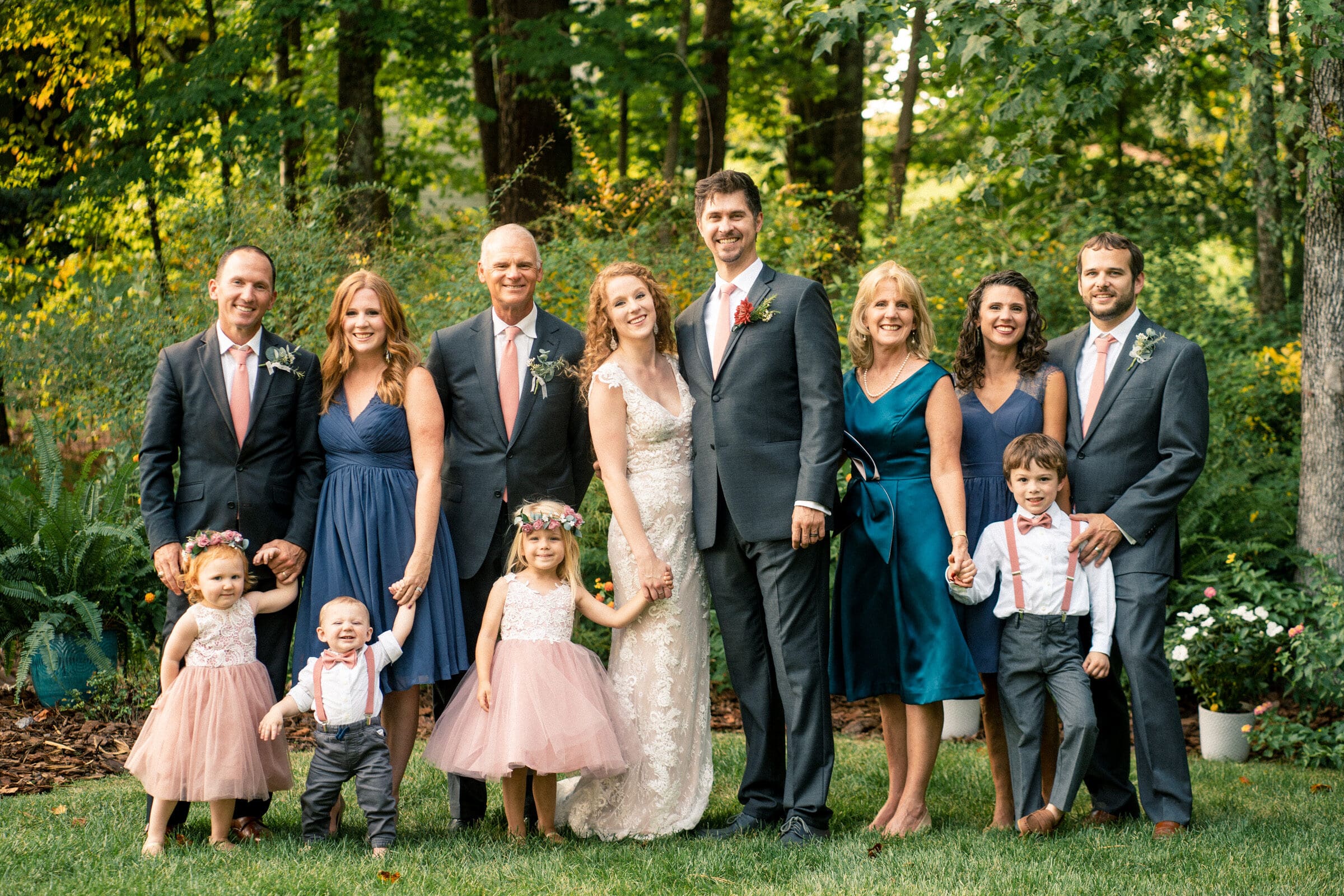 A group of adults and children dressed in formal attire pose outdoors. The adults wear suits and dresses, while the children wear white tops with pink skirts or gray pants and suspenders. They stand on a grassy area with trees in the background.