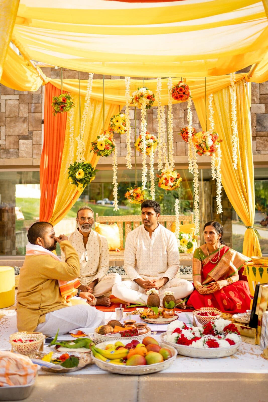A traditional ceremony takes place with vibrant yellow and orange drapes and flower decorations. Four people are seated on the floor, surrounded by fruits and ritual items, under a warm, bright setting.