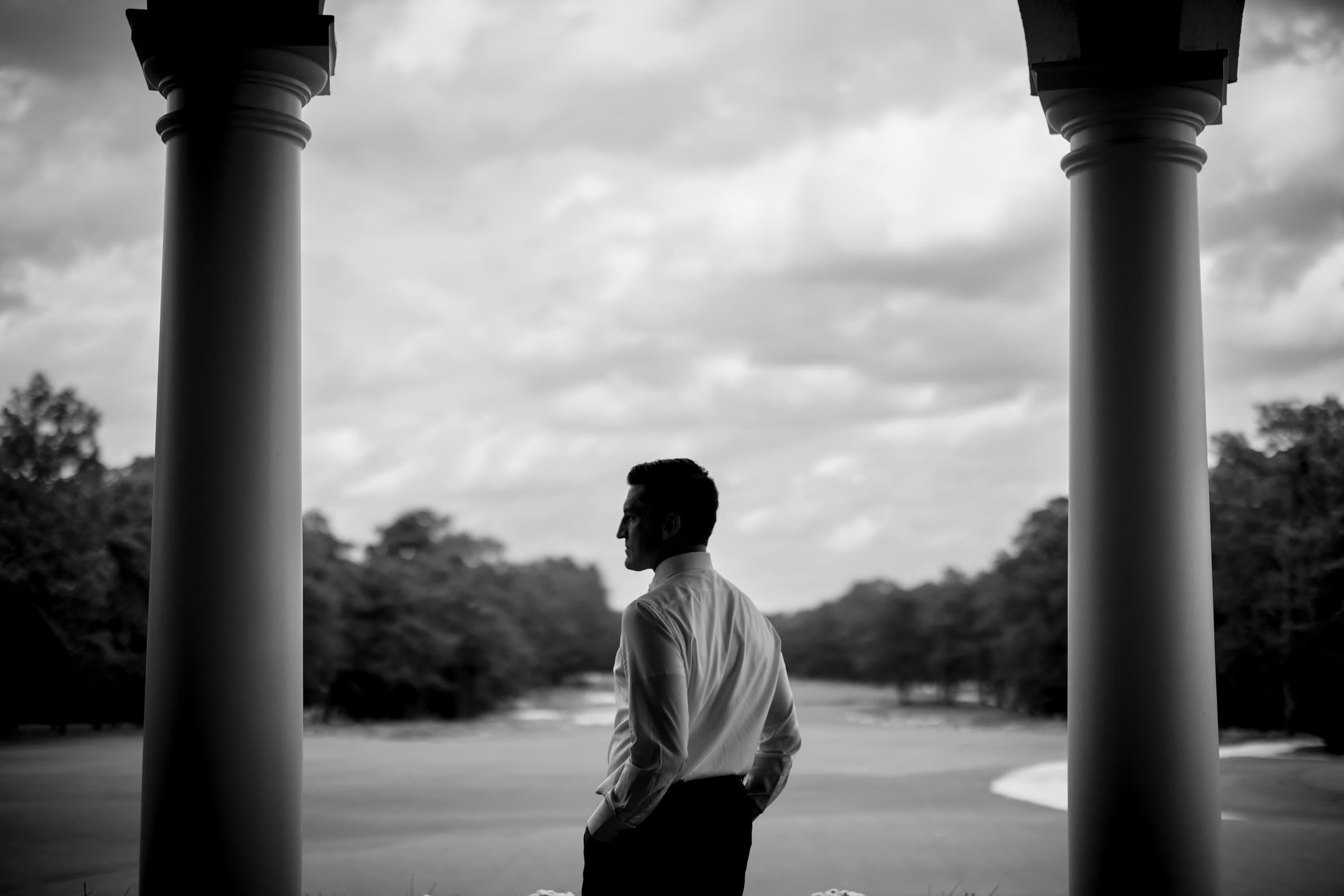 Silhouette of a person in a dress shirt standing between two columns, gazing at a vast, open landscape under a cloudy sky. The scene is captured in black and white, enhancing the serene and contemplative mood.