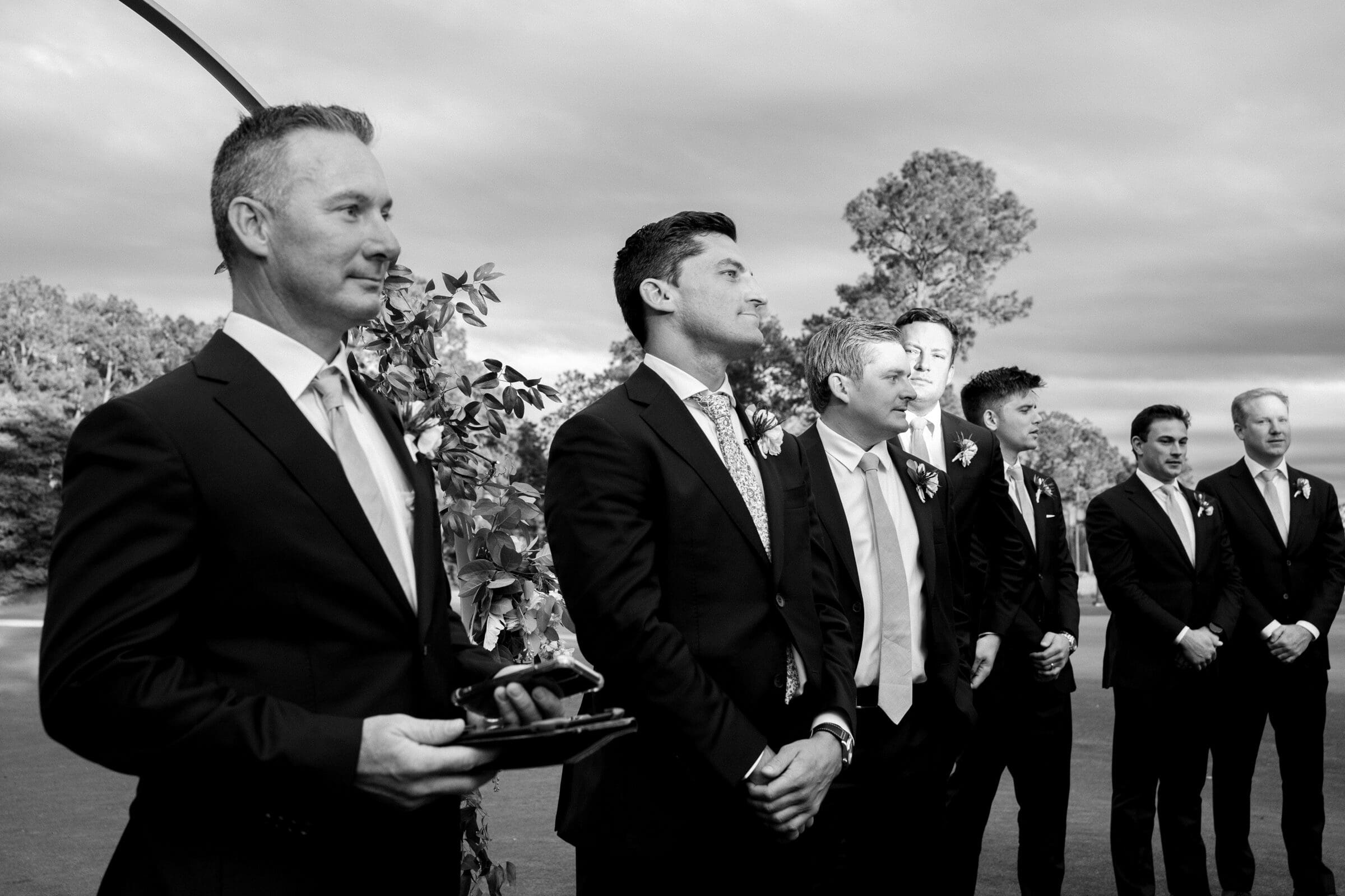 pinehurst wedding photography - lindsey & brendan A black and white photo of a wedding ceremony shows a group of men in suits standing under a floral arch. One man holds a book, possibly leading the ceremony, while others look towards the front with attentive expressions. Trees and cloudy sky in the background.