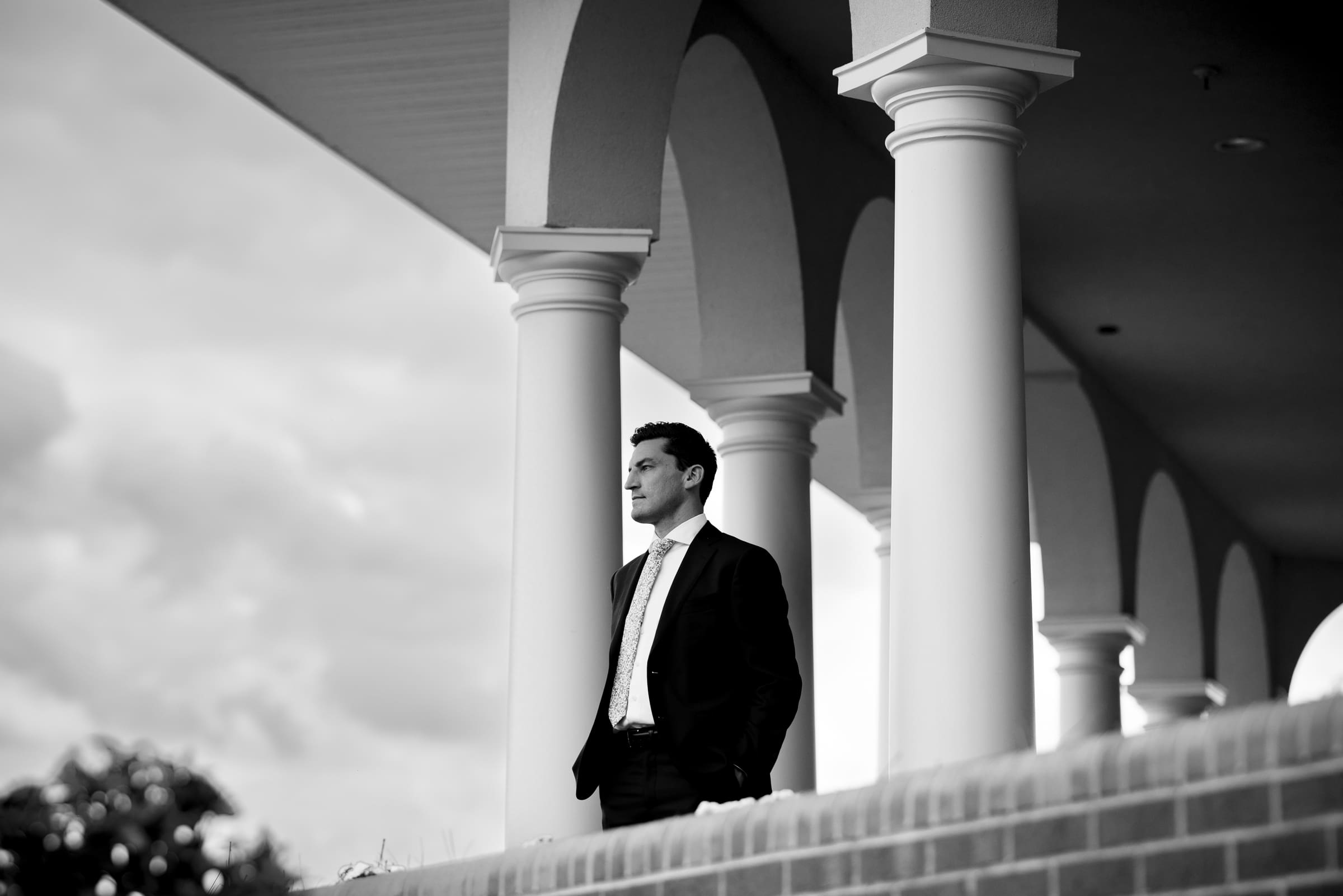pinehurst wedding photography - lindsey & brendan A black and white image of a man in a suit standing on a terrace with large columns, gazing into the distance. The sky is partly cloudy, creating a serene backdrop.
