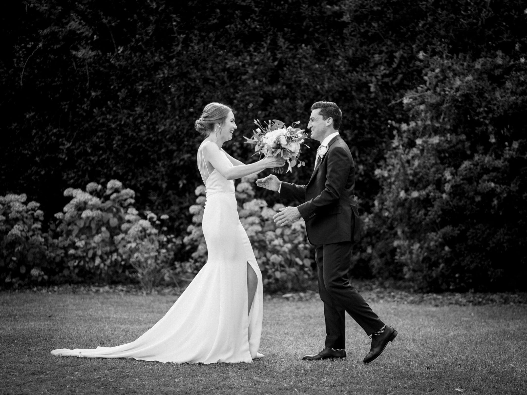 pinehurst wedding photography - lindsey & brendan A joyful couple celebrates outdoors, with the bride in a long dress with a slit and the groom in a suit. She holds a bouquet of flowers. They appear happy, surrounded by a backdrop of lush greenery and blooming plants.