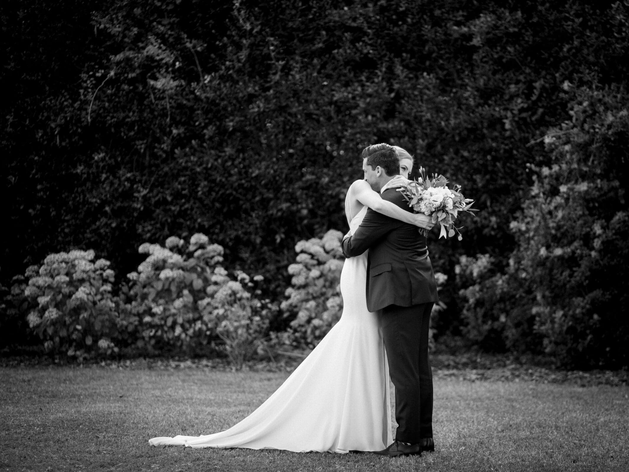pinehurst wedding photography - lindsey & brendan A black and white photo of a couple embracing outdoors. The woman is in a long, elegant dress, holding a bouquet of flowers. The man is in a suit. They stand on grass with a backdrop of dense greenery.