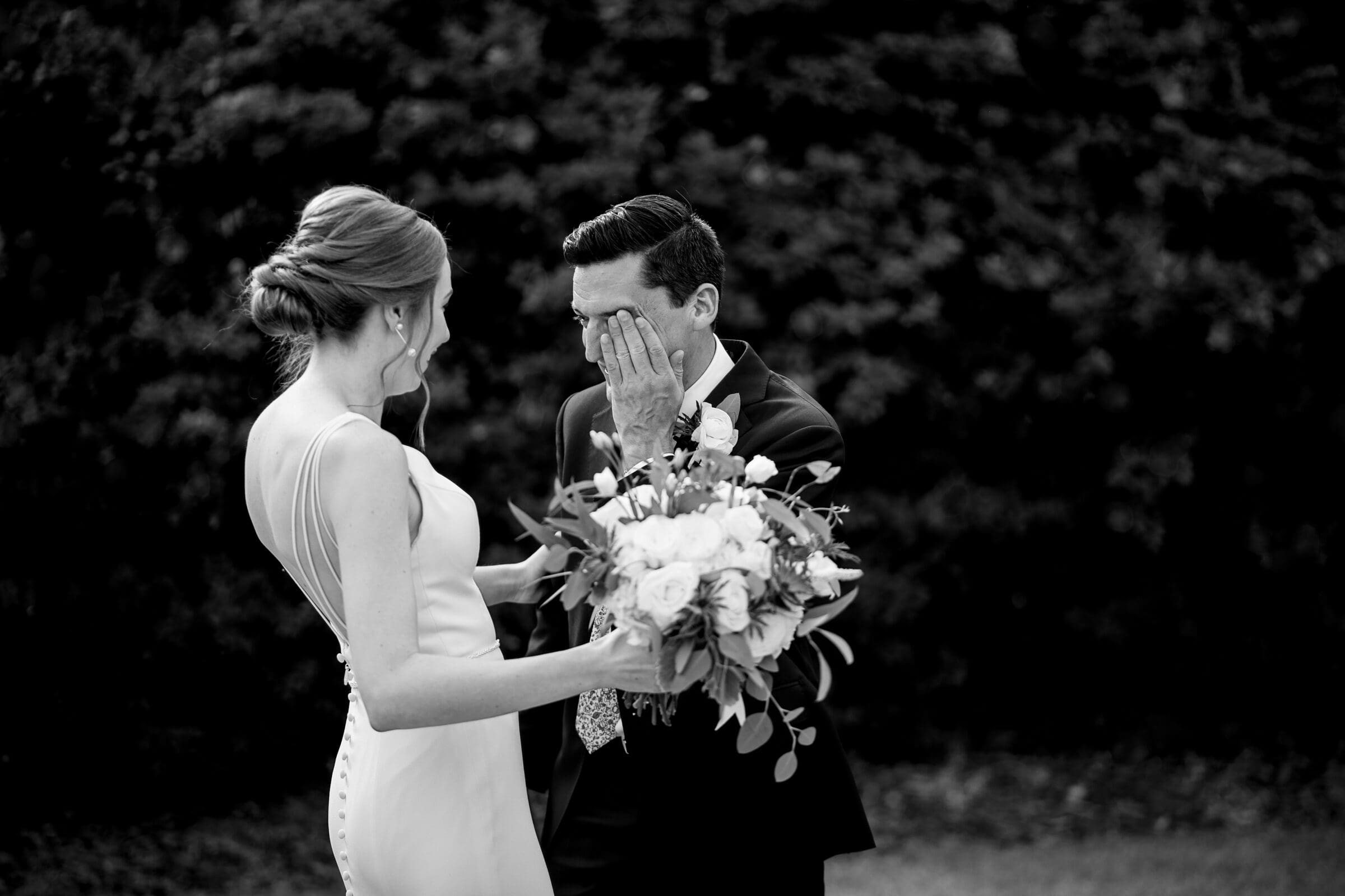 pinehurst wedding photography - lindsey & brendan A bride in a white dress holds a bouquet and smiles as a groom in a suit wipes tears from his eyes. They stand outdoors in front of a blurred backdrop of greenery. The moment is emotional and intimate.
