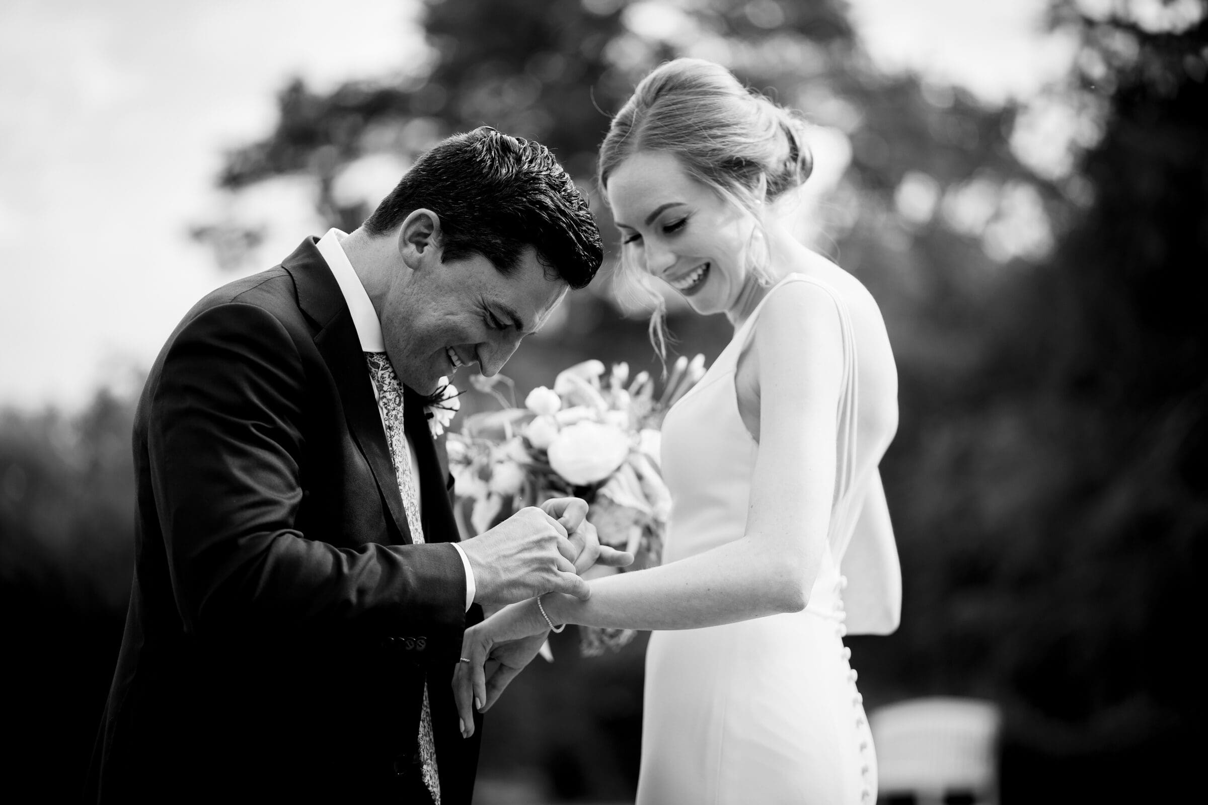 pinehurst wedding photography - lindsey & brendan A groom joyfully places a ring on the brides finger during an outdoor wedding ceremony. The bride holds a bouquet, smiling warmly. The black and white image captures a moment of happiness amid blurred natural surroundings.