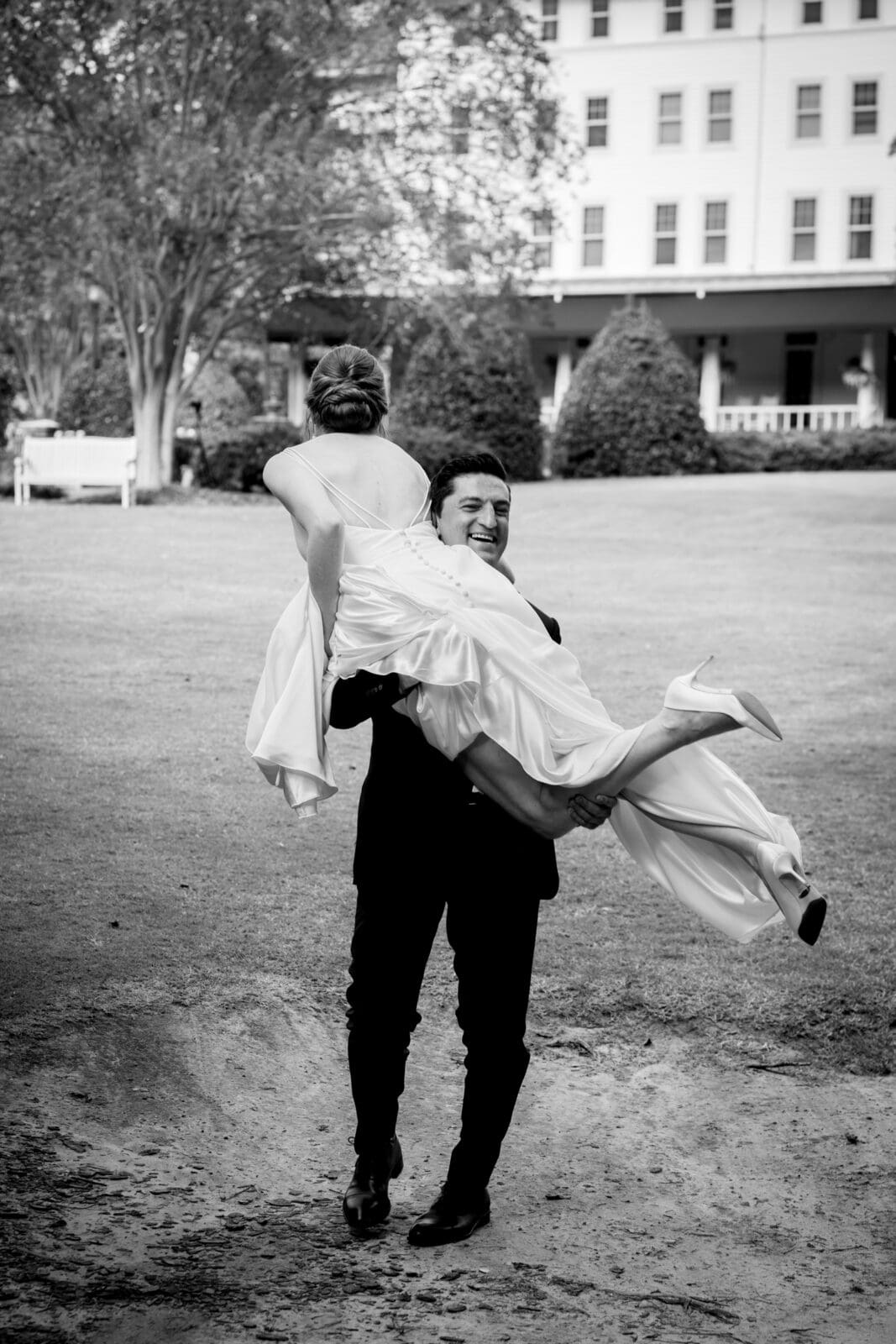 pinehurst wedding photography - lindsey & brendan A groom in a suit joyfully carries his bride, wearing a wedding dress and heels, across a grassy area. In the background, theres a large building with trees and a porch. The image is in black and white.