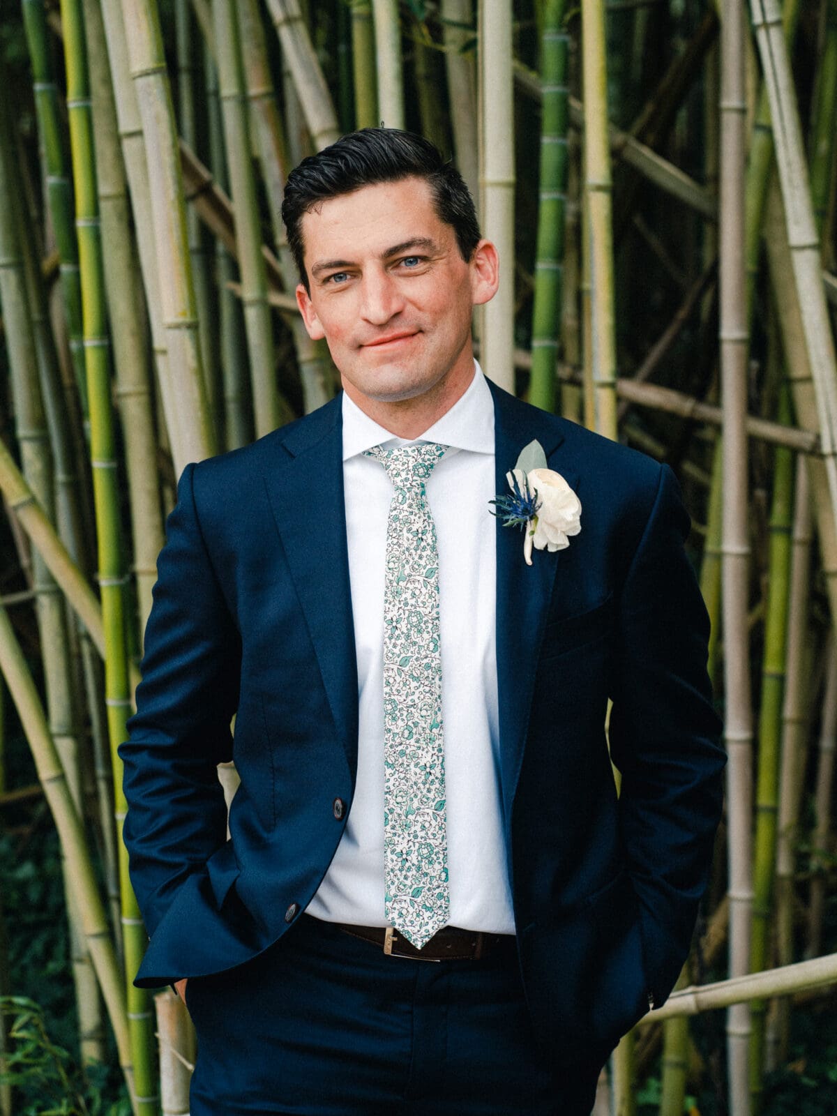 pinehurst wedding photography - lindsey & brendan A man in a navy suit and floral tie stands confidently with his hands in his pockets in front of a bamboo grove. He has a boutonniere on his lapel and a slight smile.