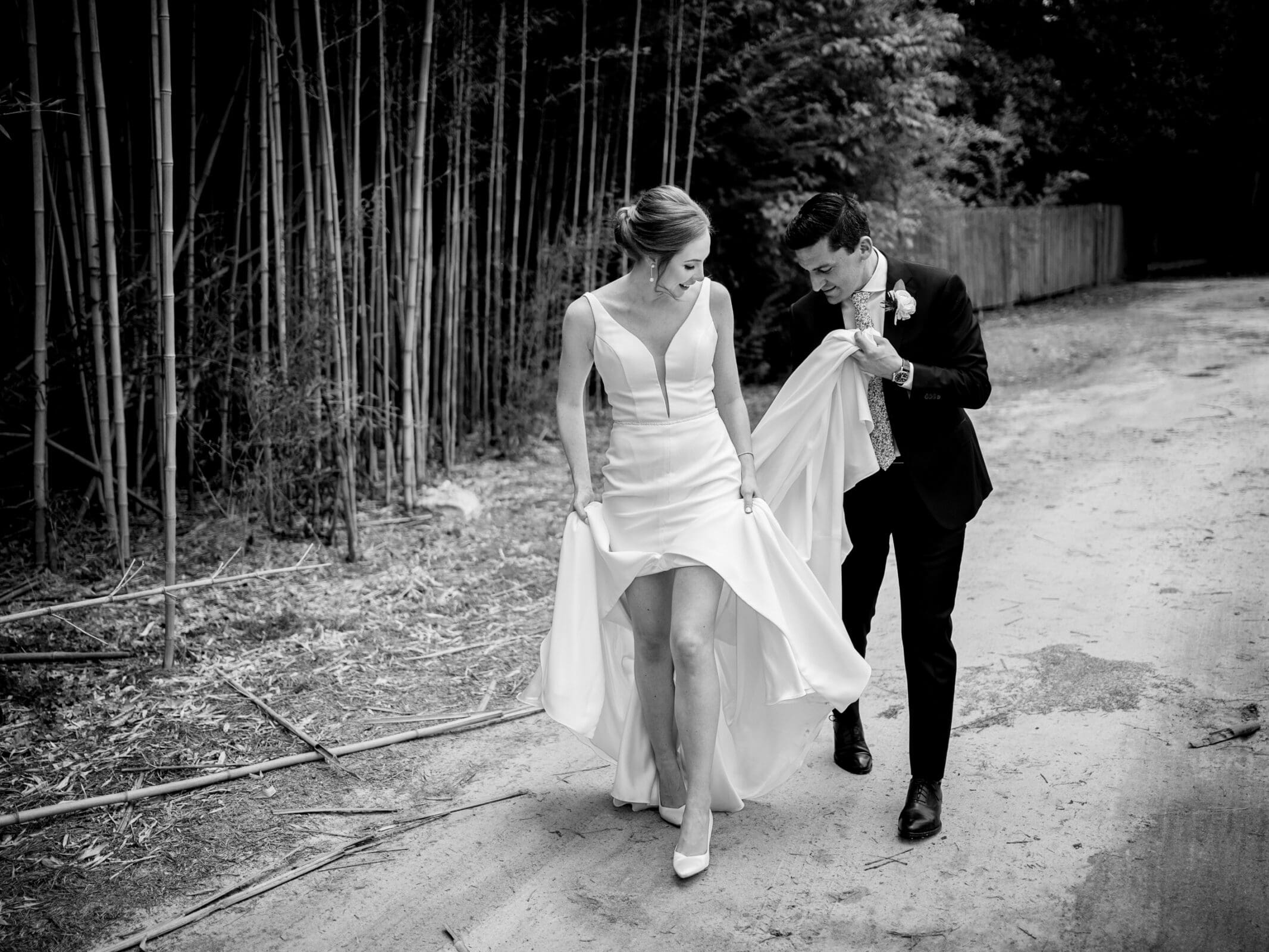 pinehurst wedding photography - lindsey & brendan A bride in a white dress and a groom in a black suit walk along a path lined with bamboo. The groom holds the brides train, and they both look down as they walk, smiling. The image is in black and white.