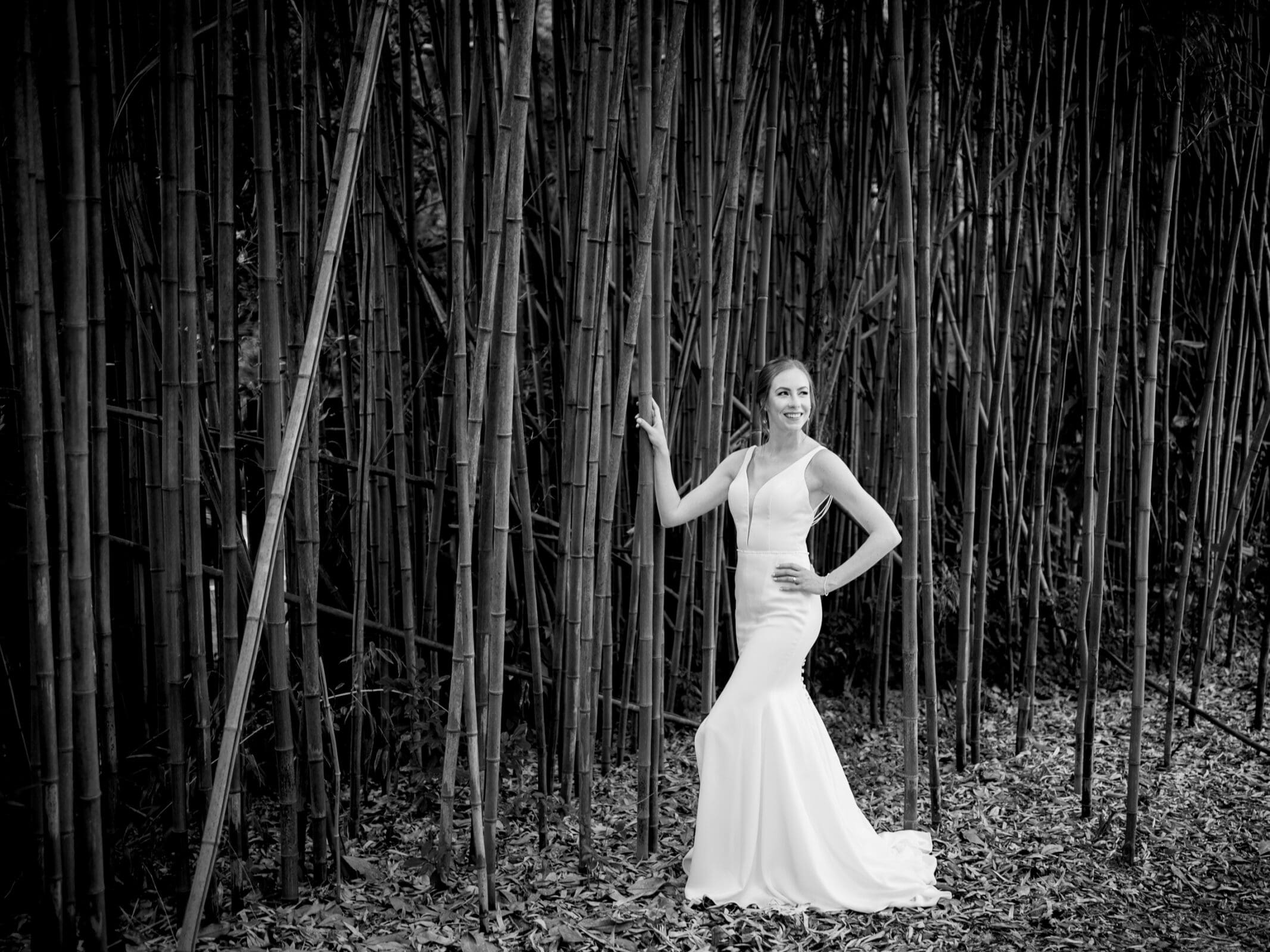 pinehurst wedding photography - lindsey & brendan A woman in a white wedding dress poses confidently against a backdrop of tall bamboo stalks. She has one hand on her hip and the other holding a bamboo stalk. The ground is covered with fallen leaves. The image is in black and white.