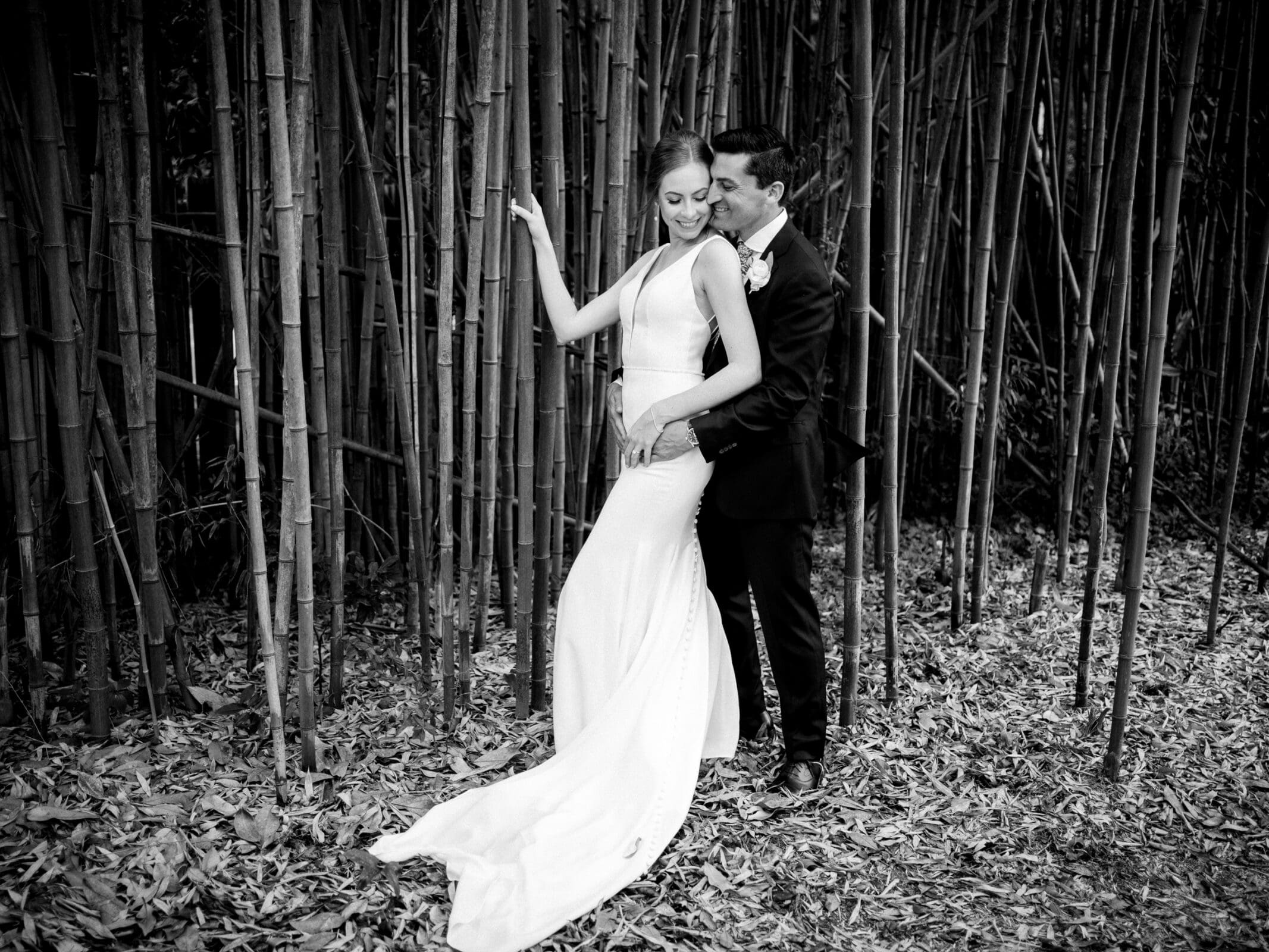 pinehurst wedding photography - lindsey & brendan A bride in a long, sleeveless gown and a groom in a dark suit stand embraced in front of tall bamboo stalks. The ground is covered with dry leaves. The groom lovingly kisses the brides cheek as she smiles, holding her bouquet.