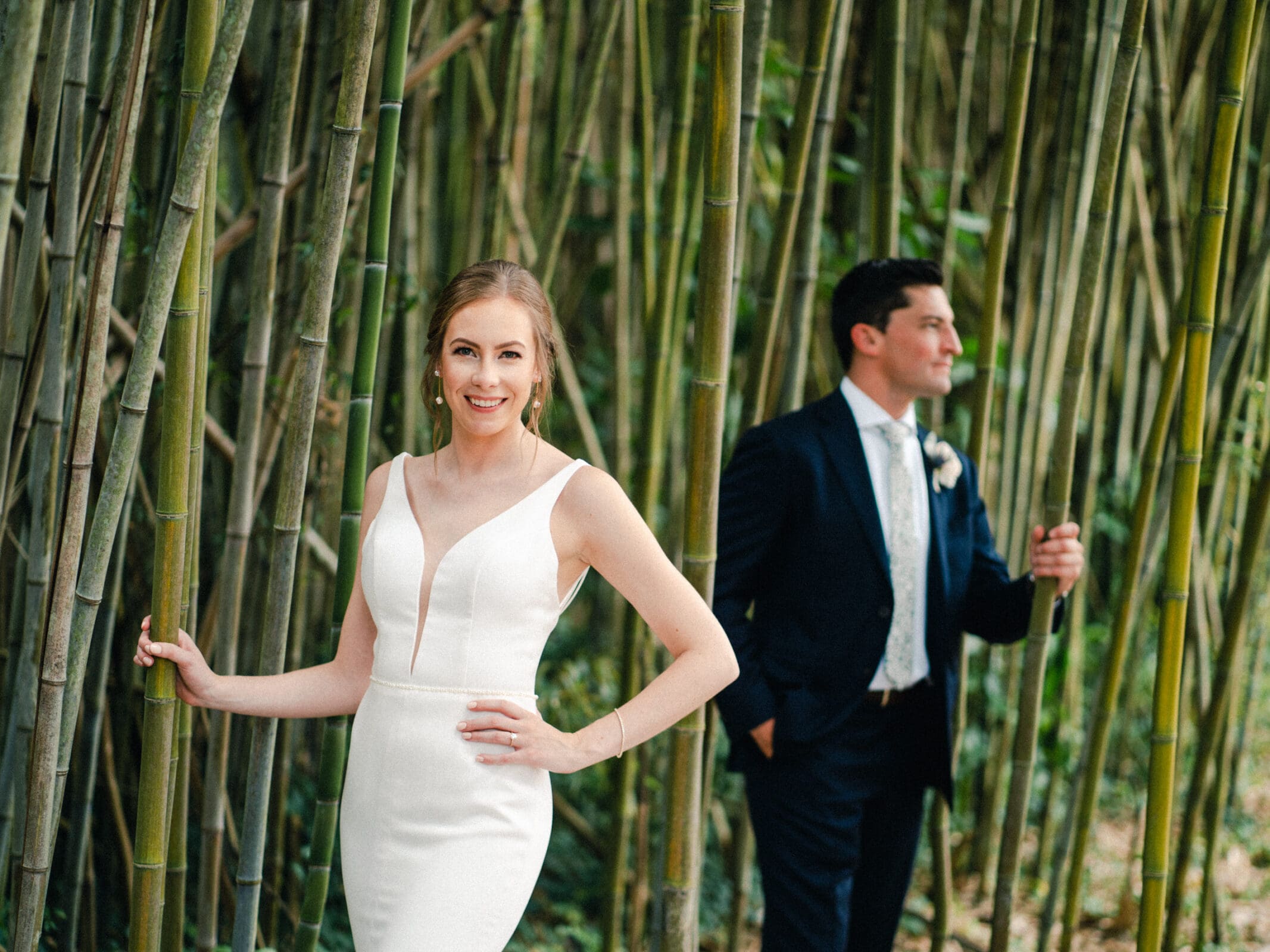 pinehurst wedding photography - lindsey & brendan A bride in a white dress smiles while standing among tall bamboo stalks. A groom in a dark suit stands slightly behind her, leaning on a bamboo stalk. They are outdoors, surrounded by lush greenery.