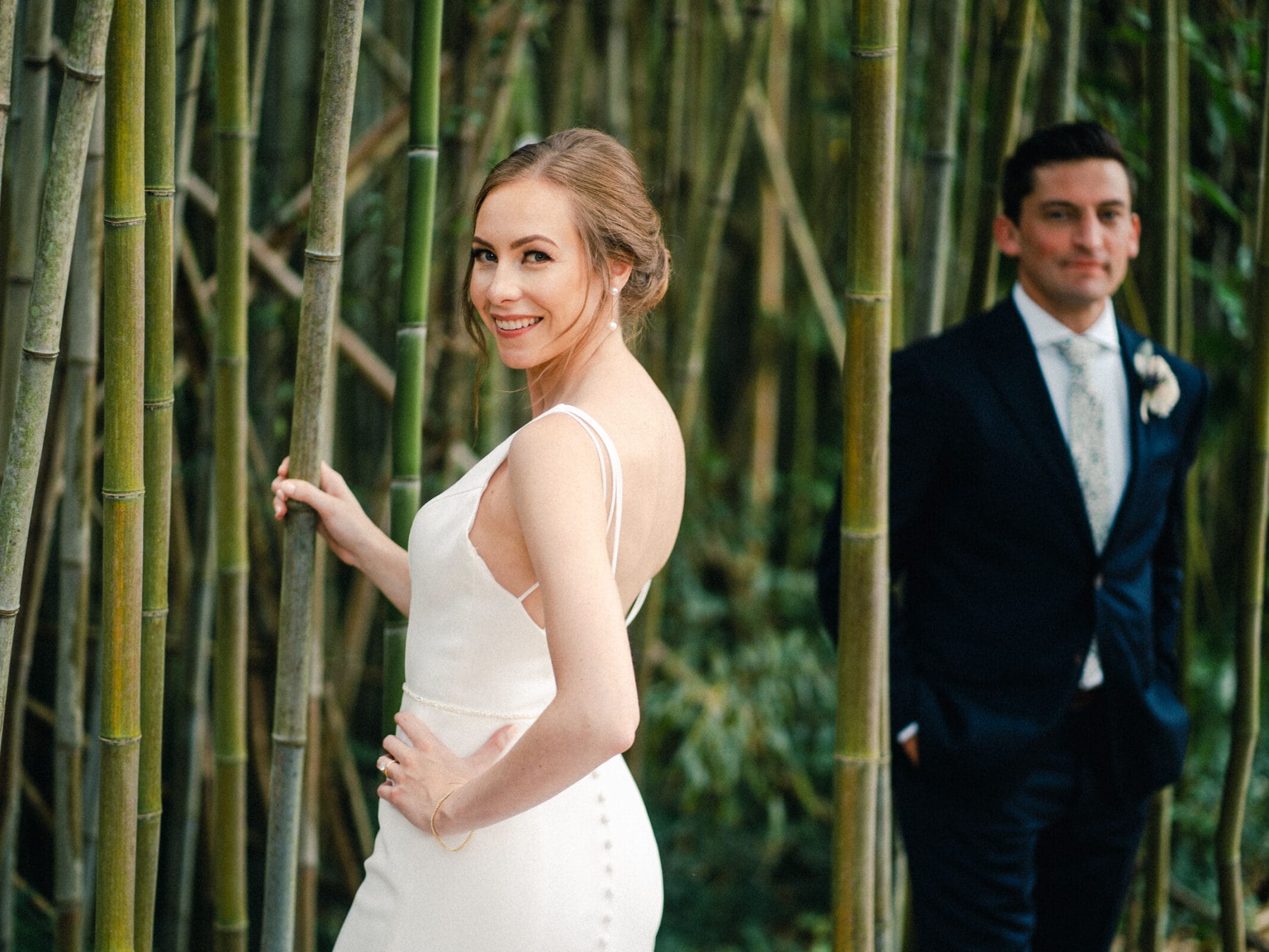 A bride smiles at the camera while standing in a bamboo grove, wearing a white dress. Behind her, a groom in a dark suit stands slightly out of focus. The setting is lush and green, creating a serene and natural backdrop.