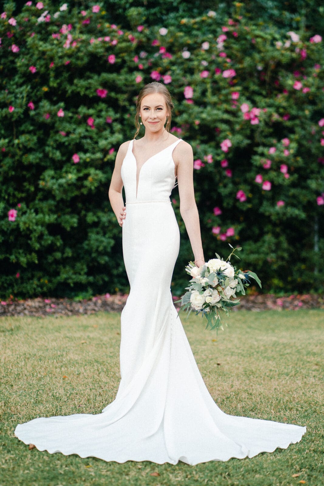 pinehurst wedding photography - lindsey & brendan A bride in a fitted white wedding dress stands on grass, holding a bouquet of white flowers. She smiles in front of a lush green hedge with blooming pink flowers.