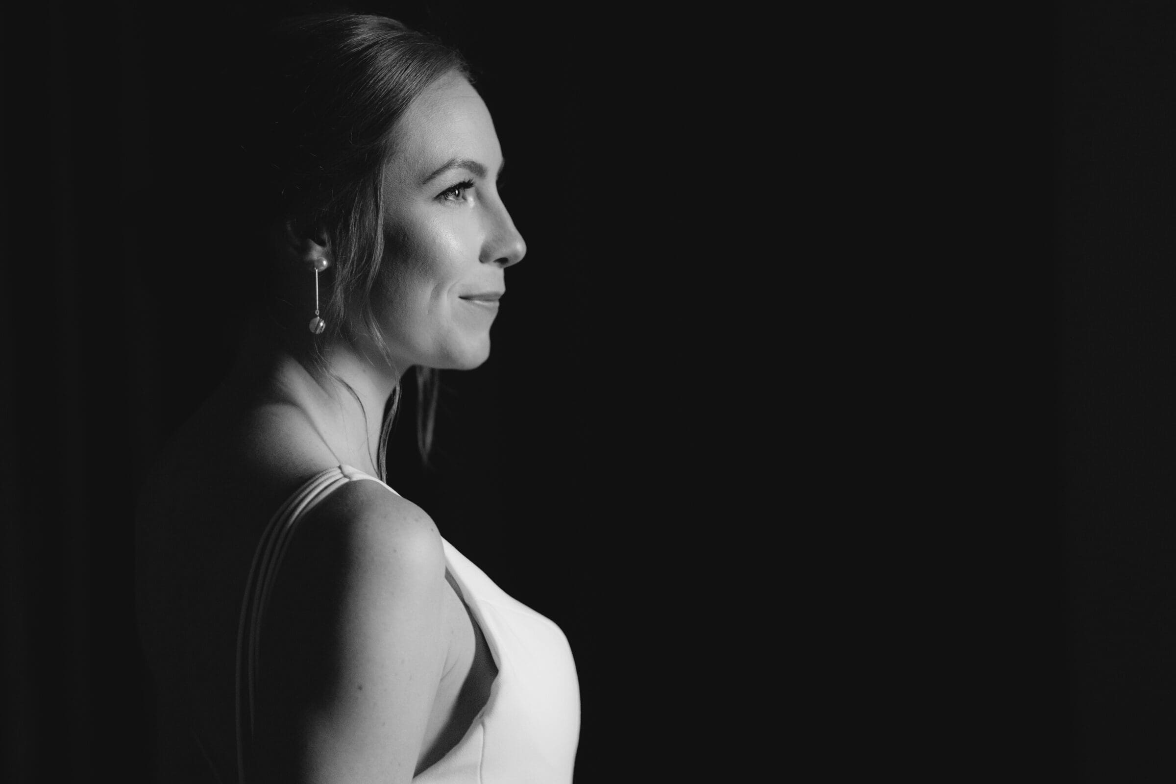 pinehurst wedding photography - lindsey & brendan Black and white portrait of a woman in profile, wearing a sleeveless dress and earrings, gazing towards the light. The background is dark, emphasizing her serene expression and softly illuminated features.