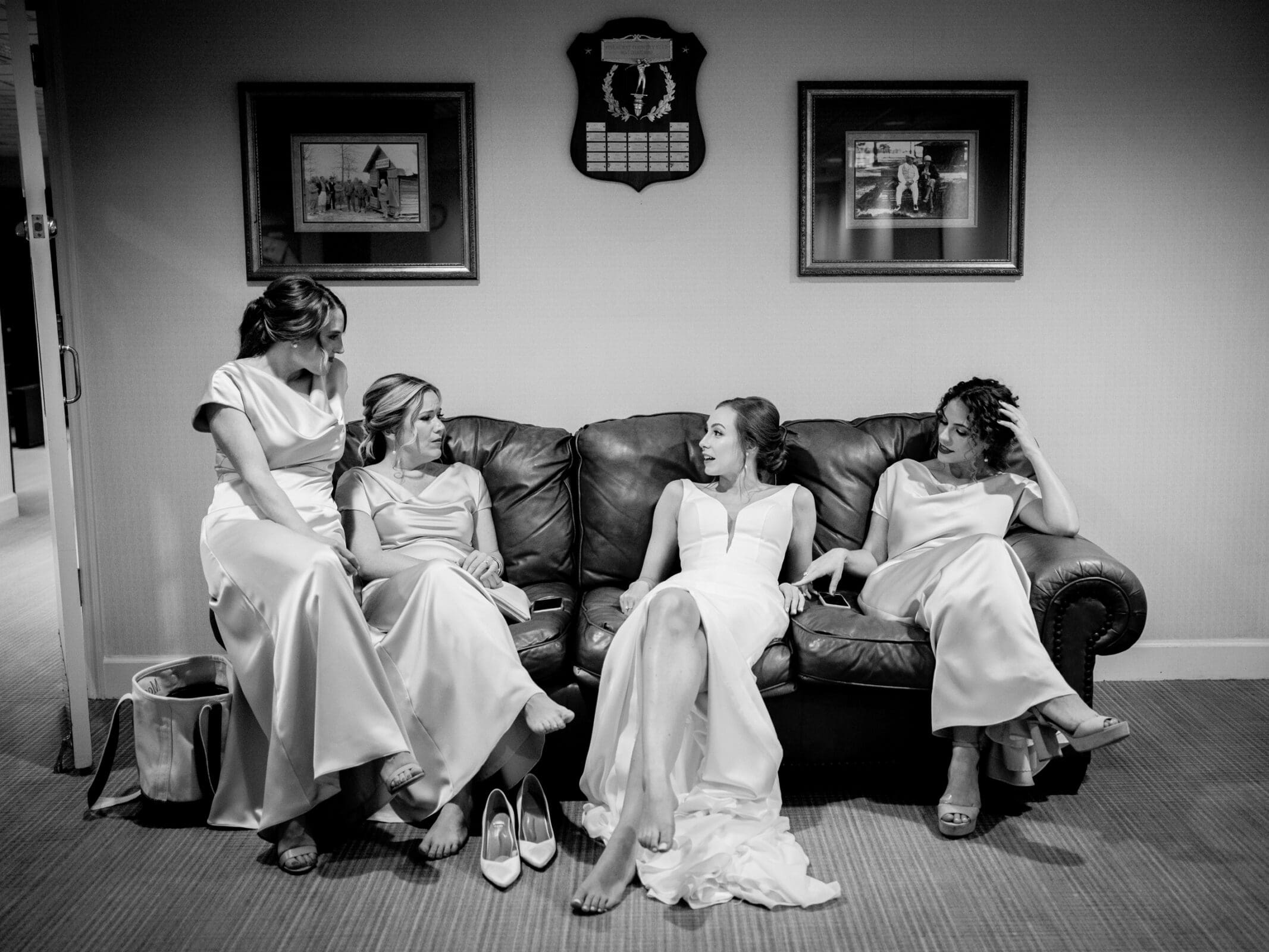 pinehurst wedding photography - lindsey & brendan Four women in elegant dresses sit on a couch in a relaxed manner. They are barefoot and appear to be sharing a light moment. Two framed pictures and a plaque are on the wall behind them. The setting is cozy and informal.