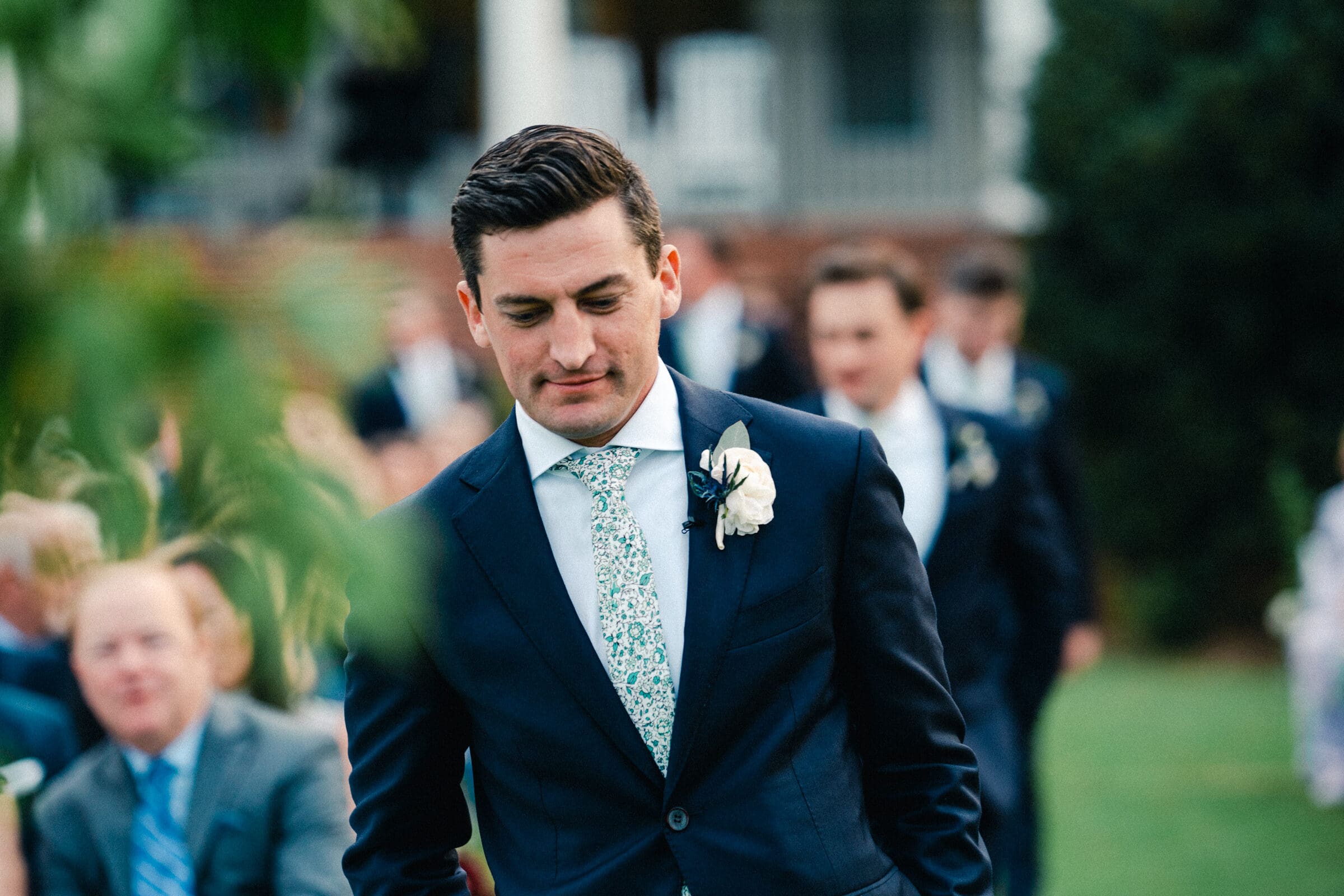 pinehurst wedding photography - lindsey & brendan A man in a navy suit with a floral tie and boutonniere walks outdoors, appearing thoughtful. A blurred background shows other suited individuals, greenery, and guests seated, suggesting a formal event.